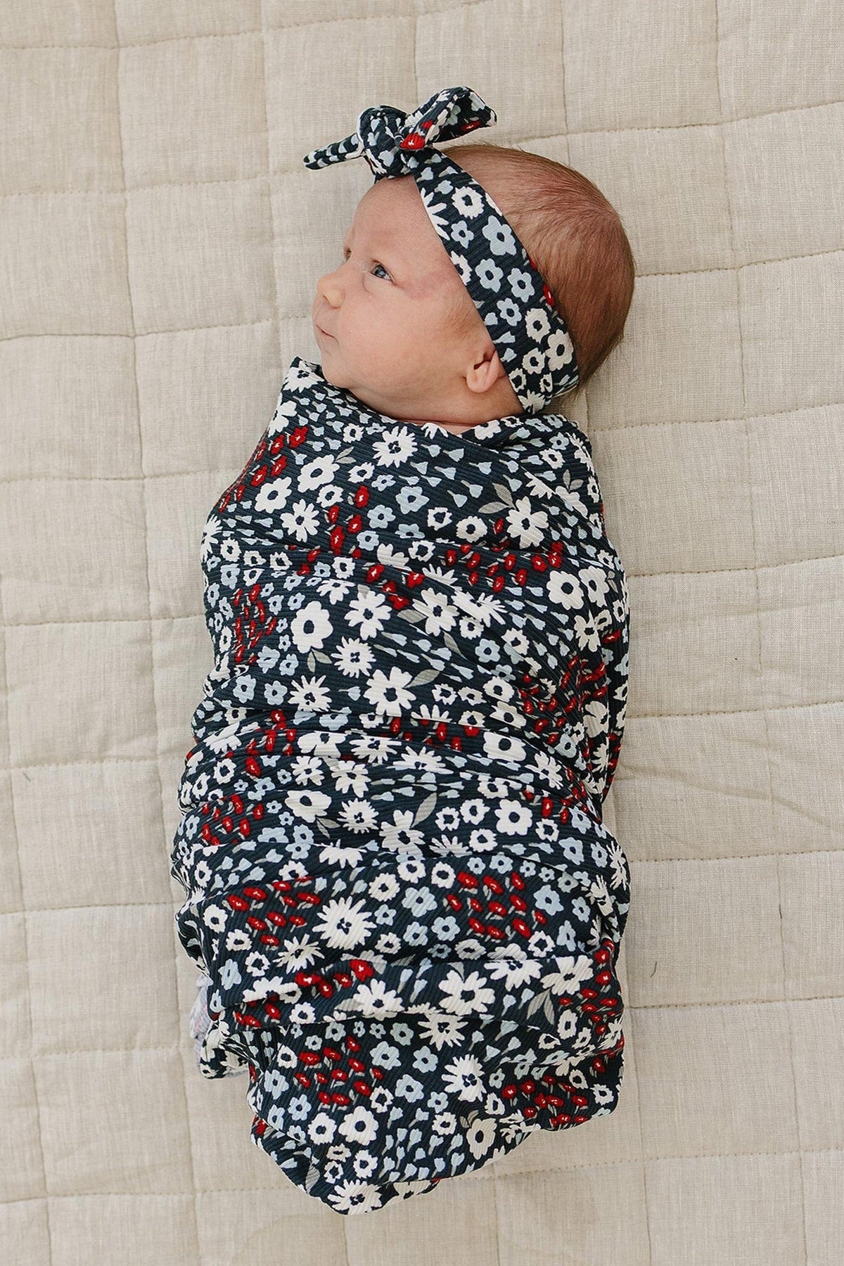 A baby wrapped in a floral-patterned swaddle with a matching headband lies on a beige quilted blanket.