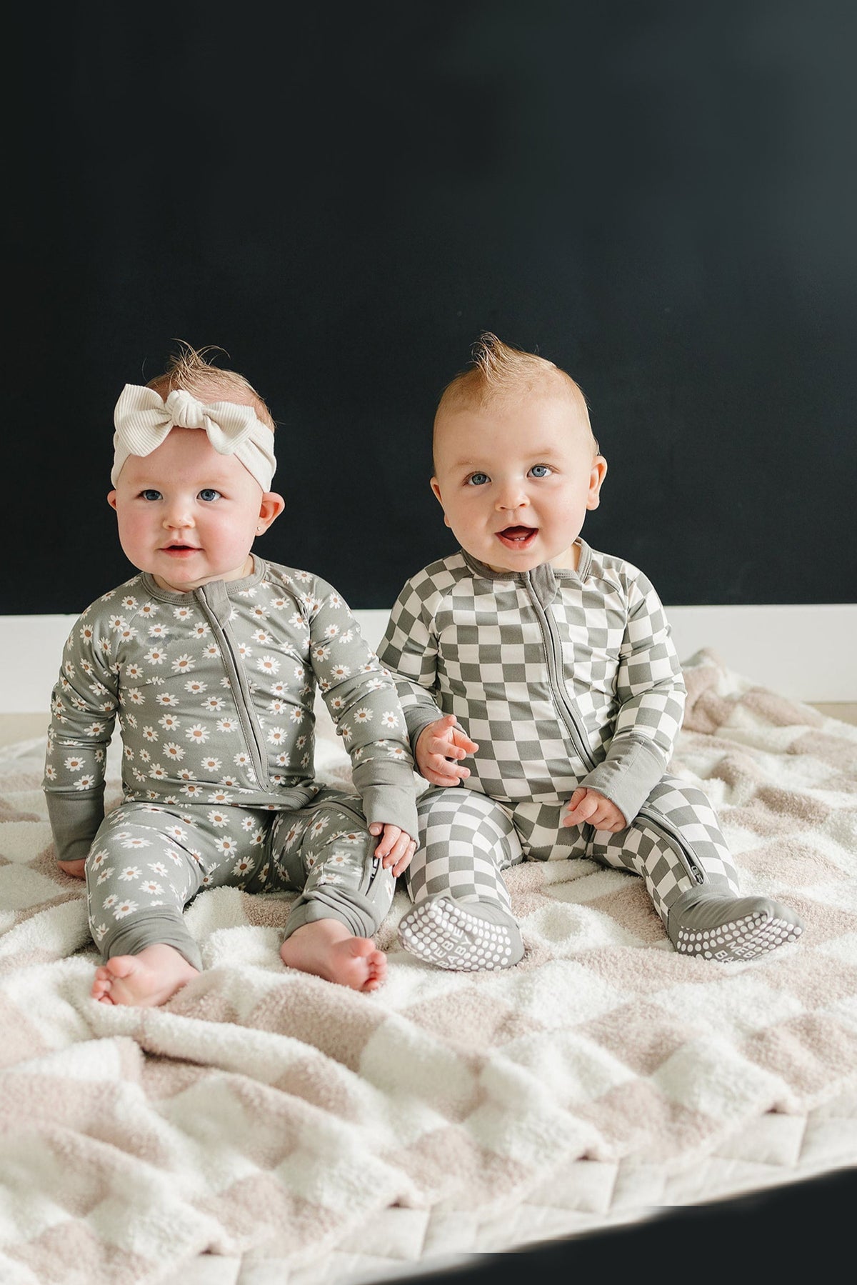 Two babies sit on a blanket, one in floral pajamas and headband, the other in checkered pajamas.