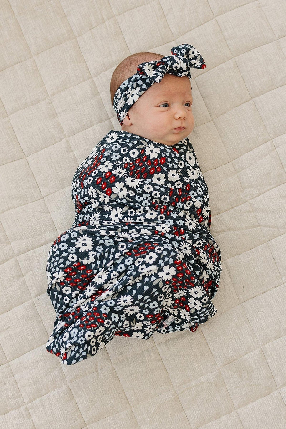 A baby wrapped in a floral-patterned swaddle and headband lies on a quilted beige blanket.