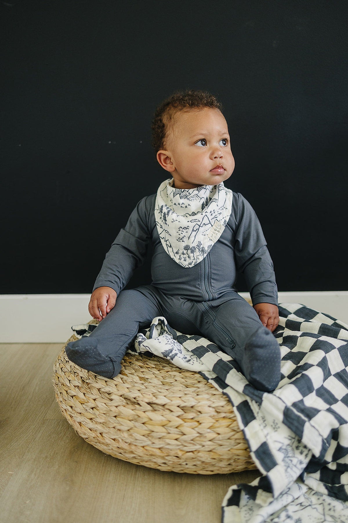 A baby wearing a mountain print muslin bib and grey outfit sits on a woven cushion with a checkered blanket.