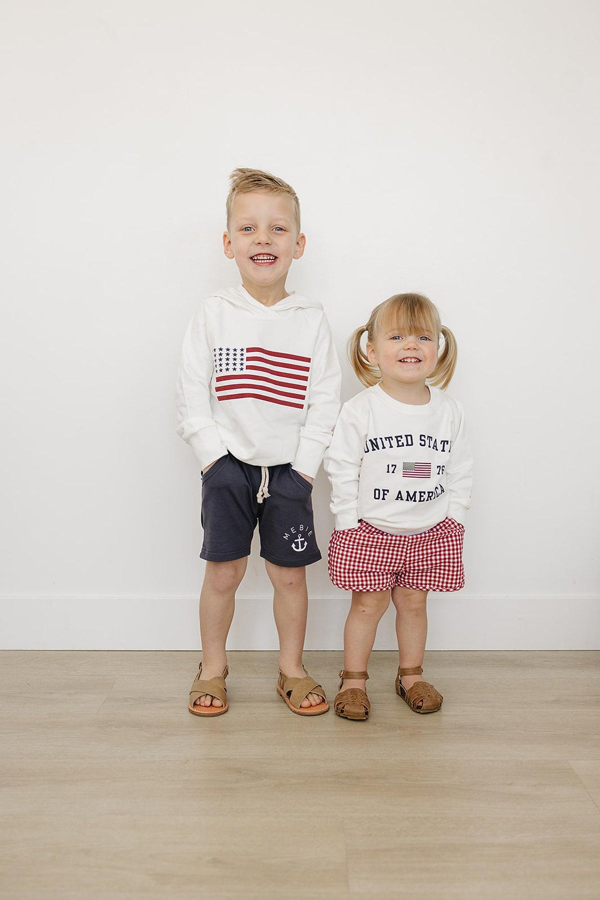 Two smiling children wearing patriotic hoodies; one with an American flag, the other with United States of America.