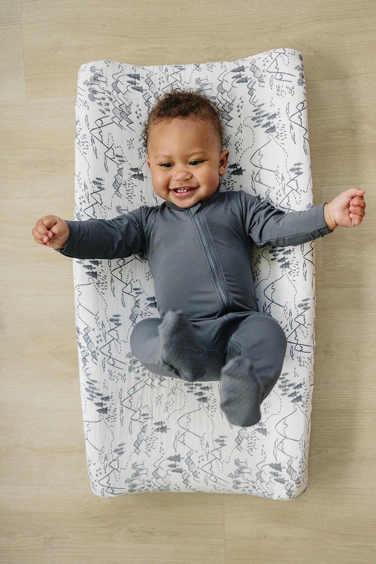 A smiling baby in a gray outfit lies on a patterned changing pad cover featuring a nature-inspired design. The cover is made from soft, breathable cotton, perfect for infants.