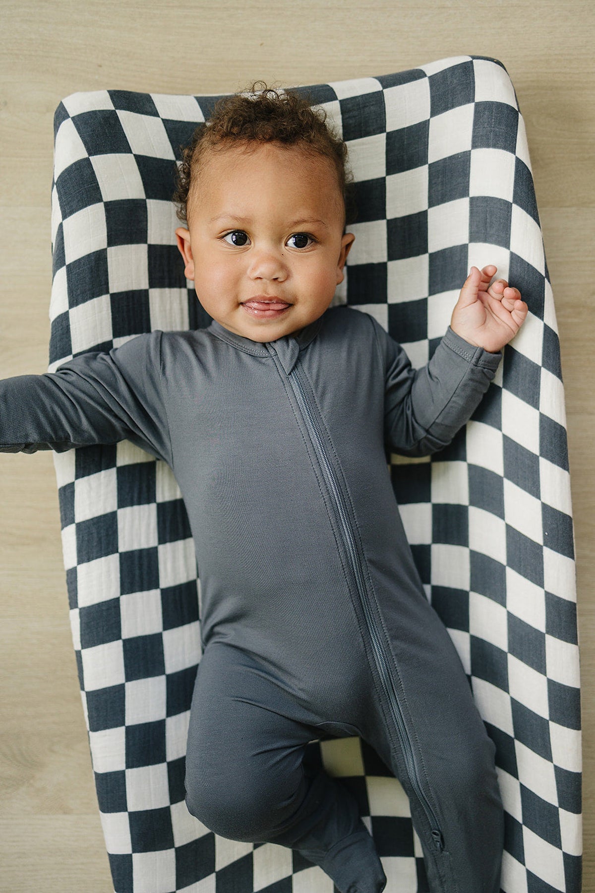 A baby in a grey onesie lies on a charcoal checkered muslin changing pad cover, looking up happily.