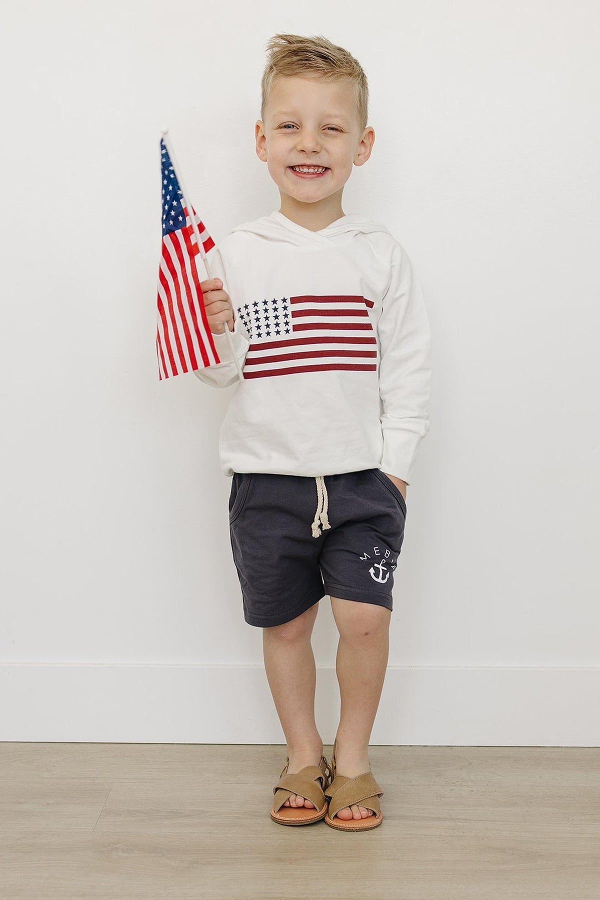 A smiling child wears a white flag hoodie and navy shorts, holding an American flag indoors.