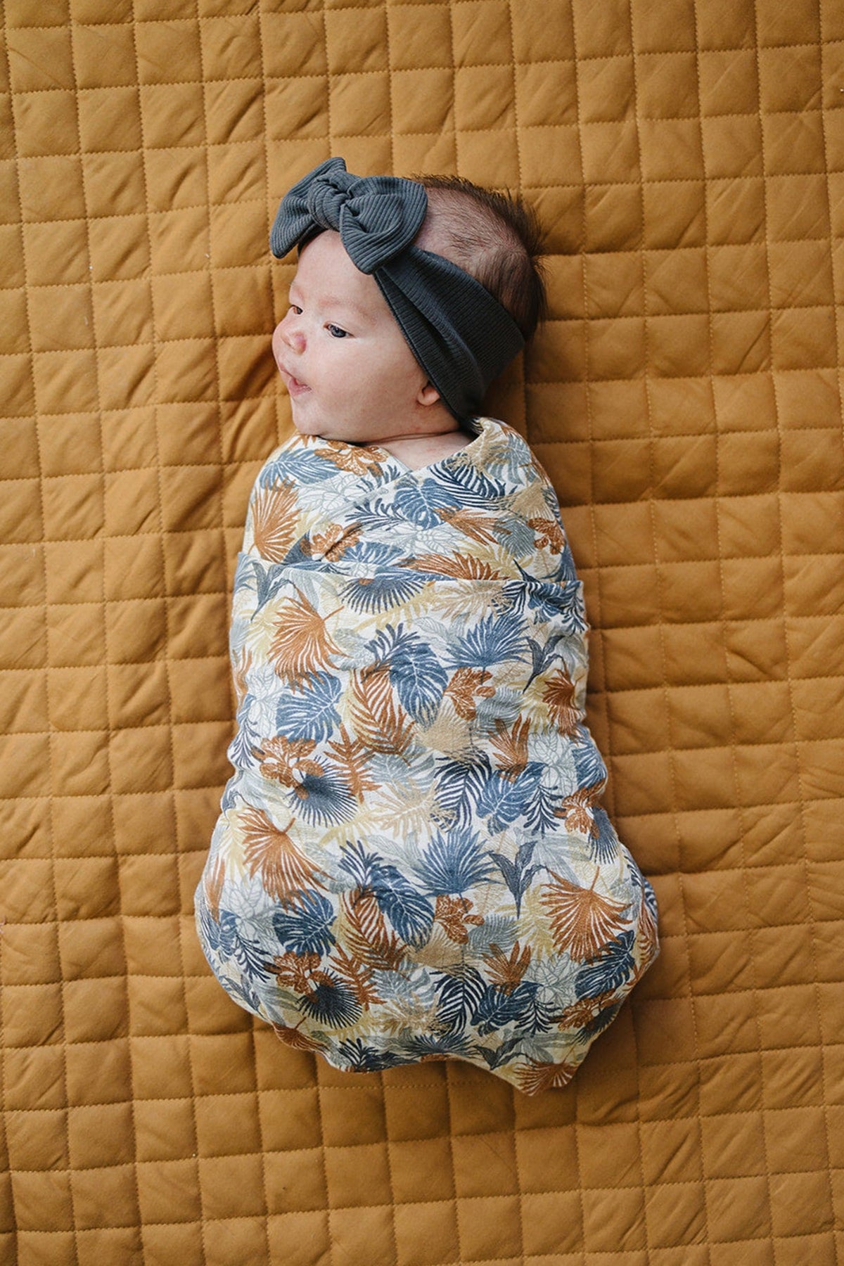 Baby wrapped in a colorful Paradise Palms muslin swaddle, lying on a quilted mustard blanket with a bow headband.
