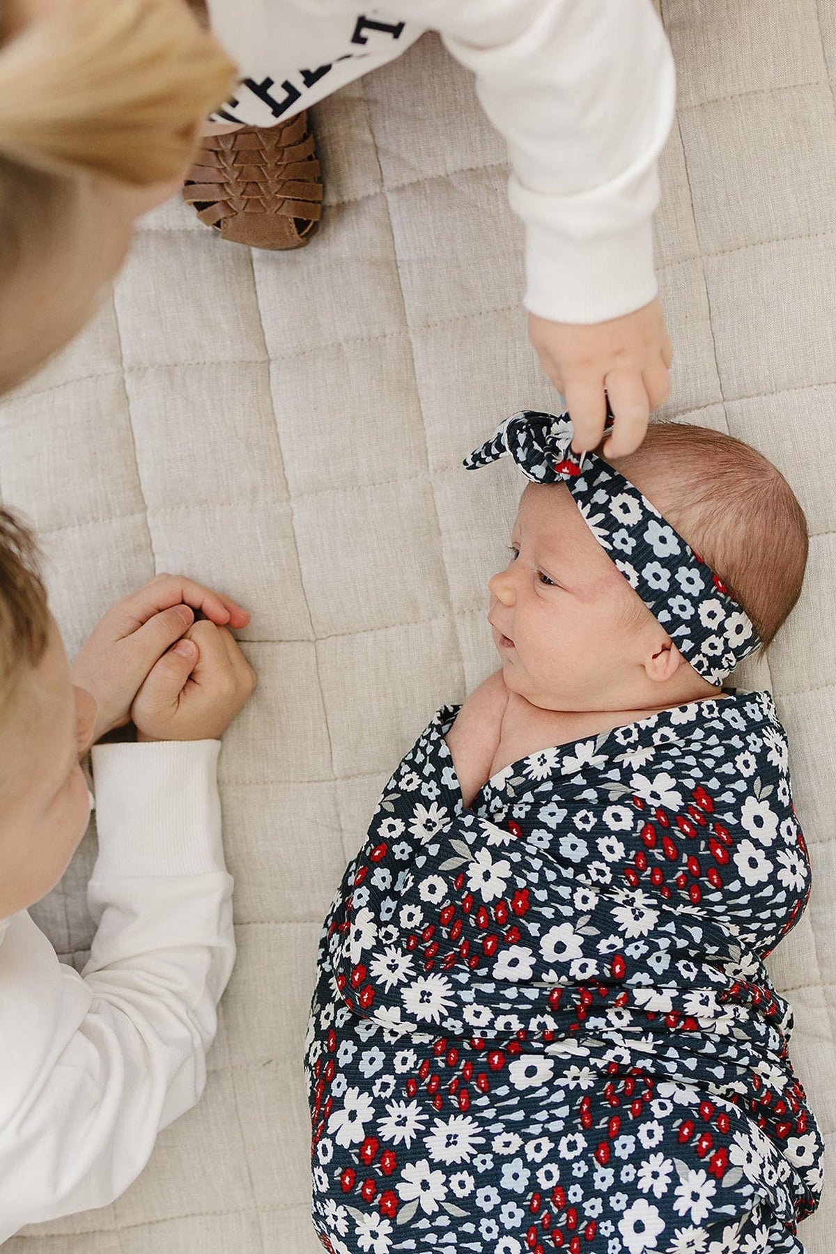 Baby wrapped in a dark floral swaddle with matching headband, lying on a quilted beige surface.