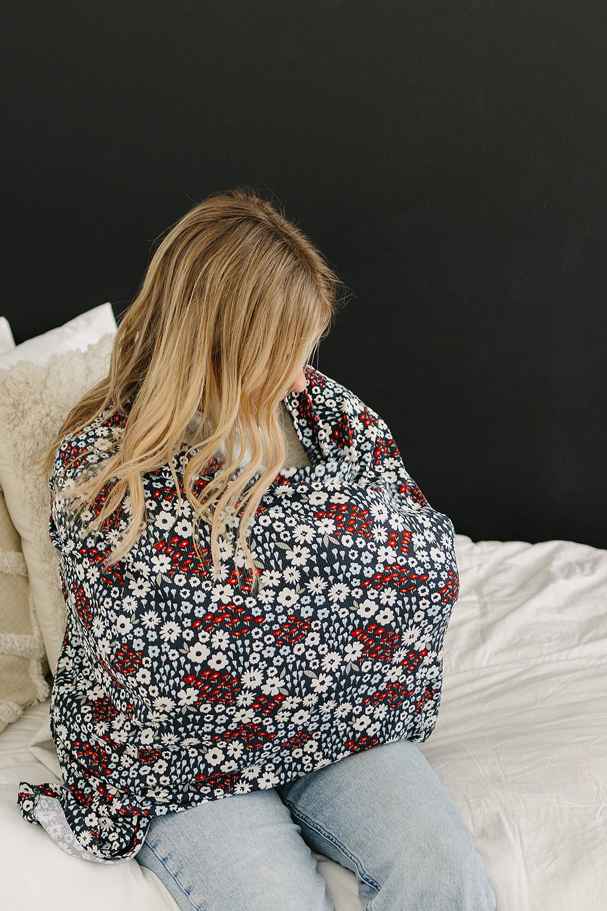 A woman sits on a bed using a floral-patterned multi-use cover with red and white flowers.