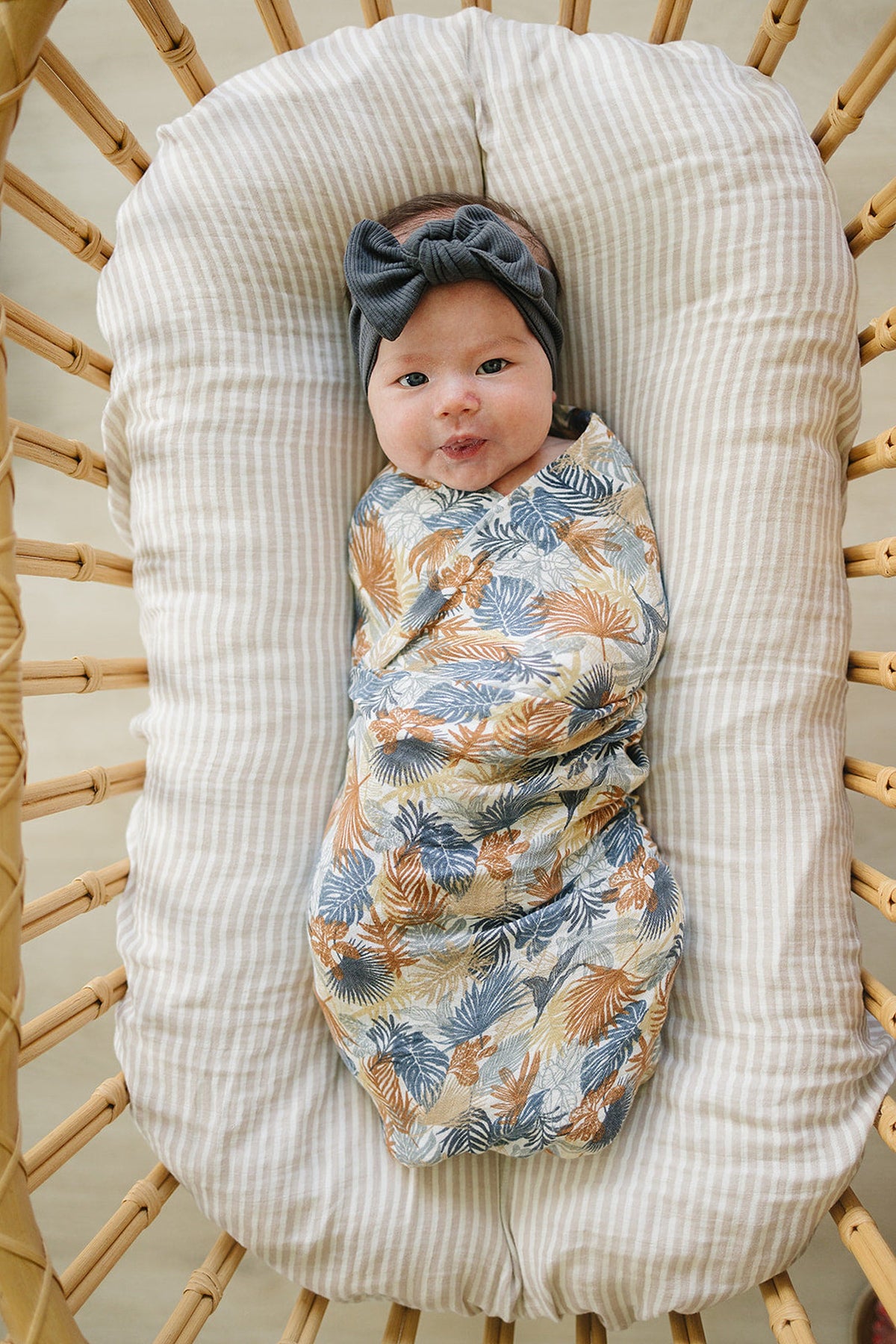 A baby wrapped in a tropical-patterned swaddle lies on a striped cushion, wearing a dark bow headband.