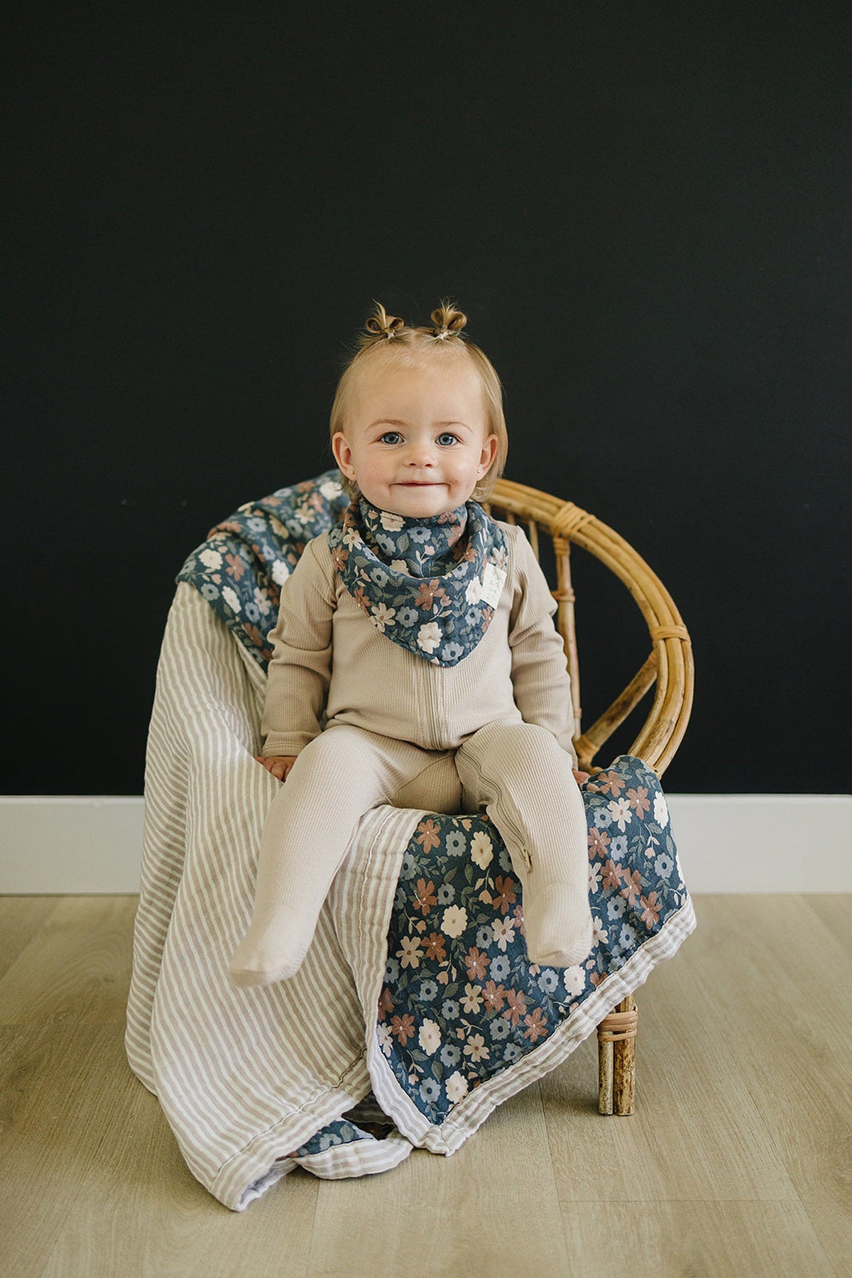 A baby wearing a floral muslin bib and beige outfit sits on a wicker chair with a matching blanket.