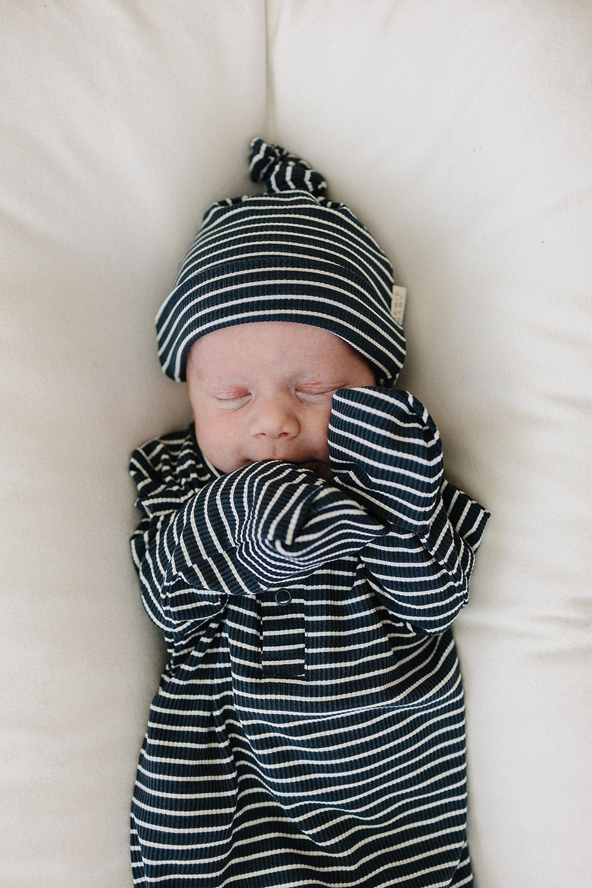 Newborn wearing a navy striped ribbed knot hat and matching gown, peacefully sleeping on a soft white surface.