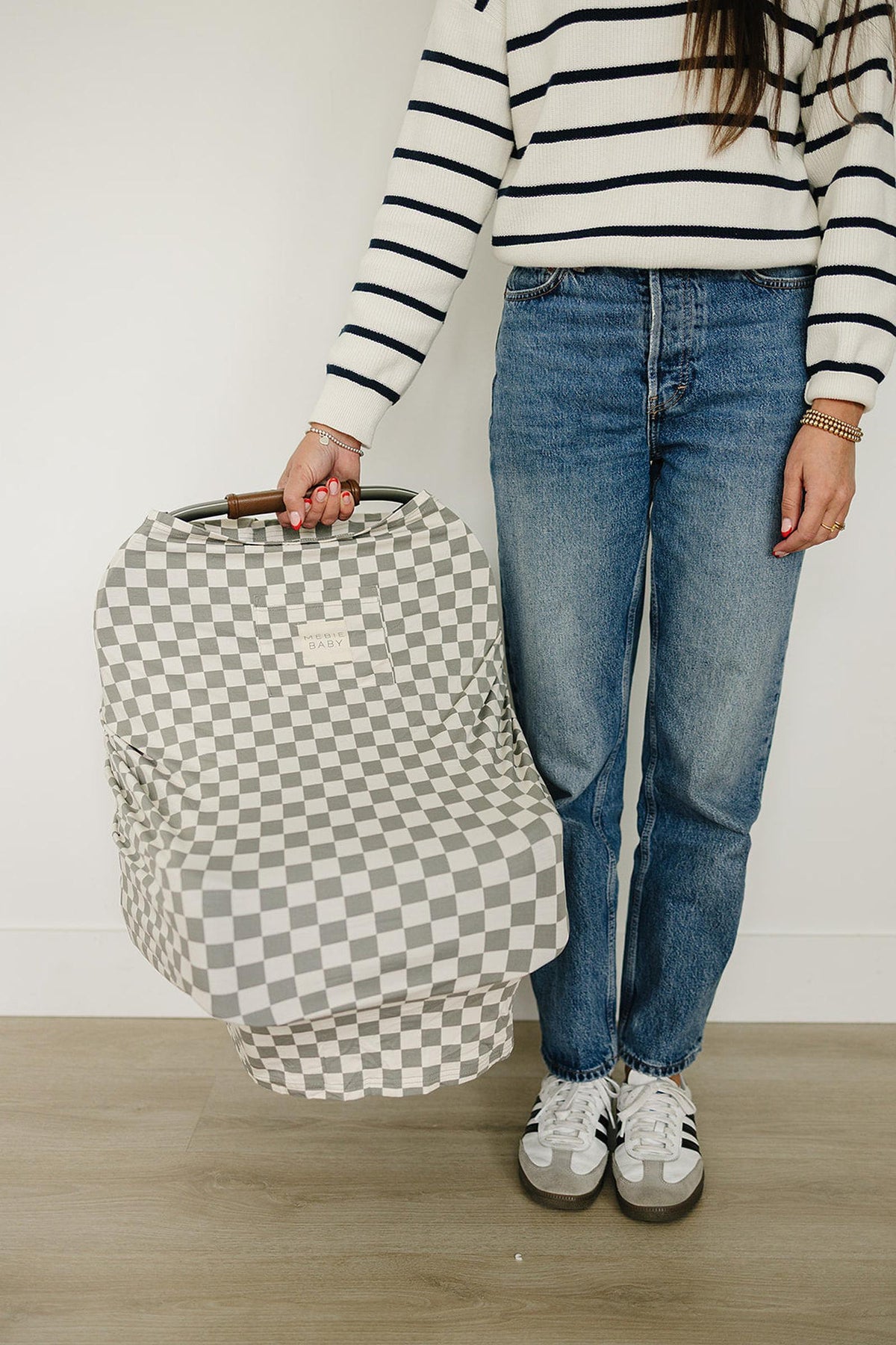 A person holds a car seat covered with a light green checkered, multi-use bamboo cover, standing on a wooden floor.