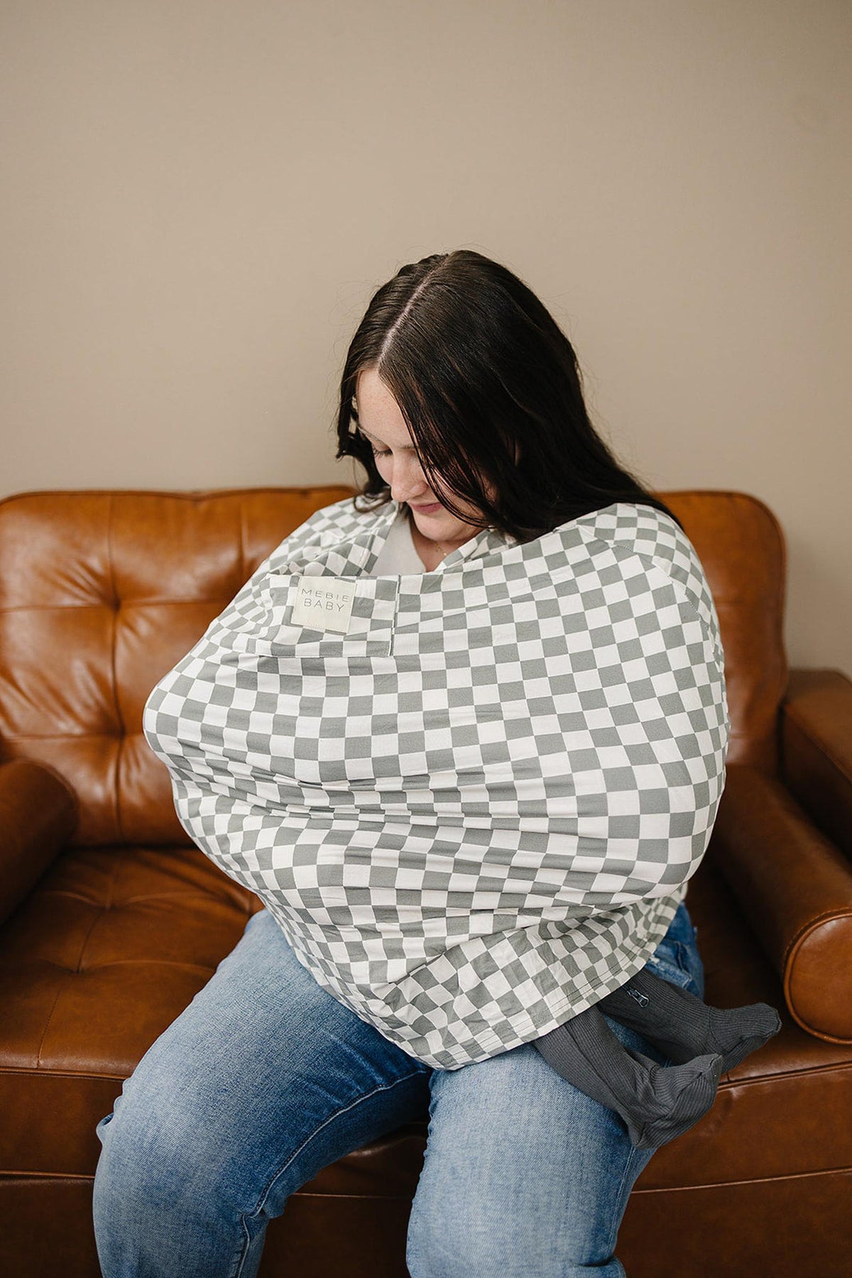 A woman sits on a brown leather couch wearing a light green checkered Mebie Baby nursing cover.