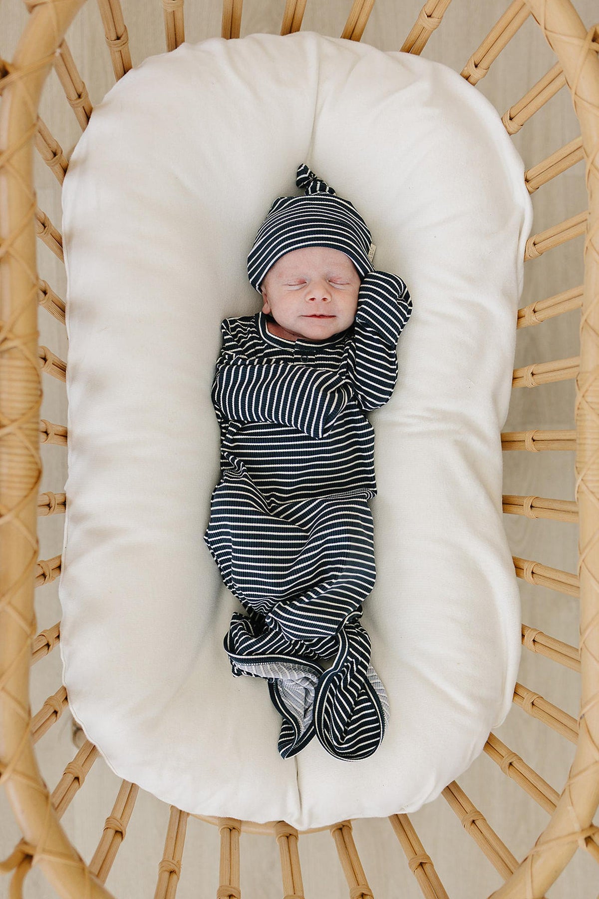 Newborn in navy striped knot gown and hat, sleeping in a wicker bassinet with a white cushion.