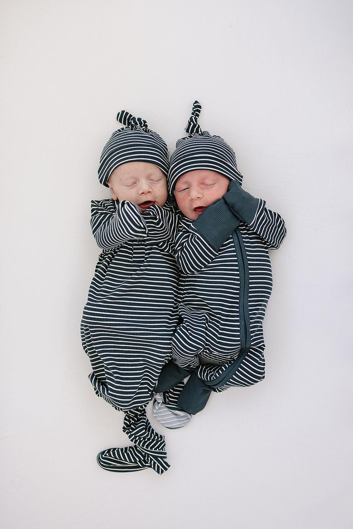 Two newborns wearing navy striped pajamas with knotted hats lie asleep on a white surface.