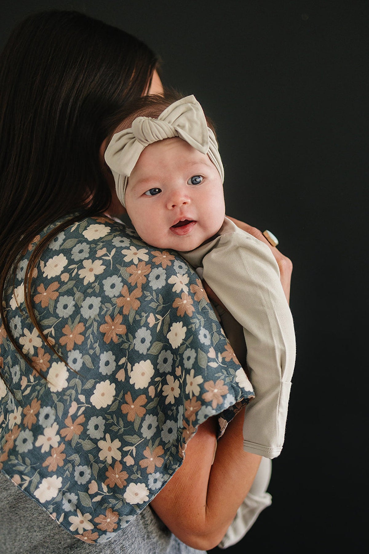 A mother holds her baby, who wears a light-colored headband, against a backdrop of a floral-patterned fabric.