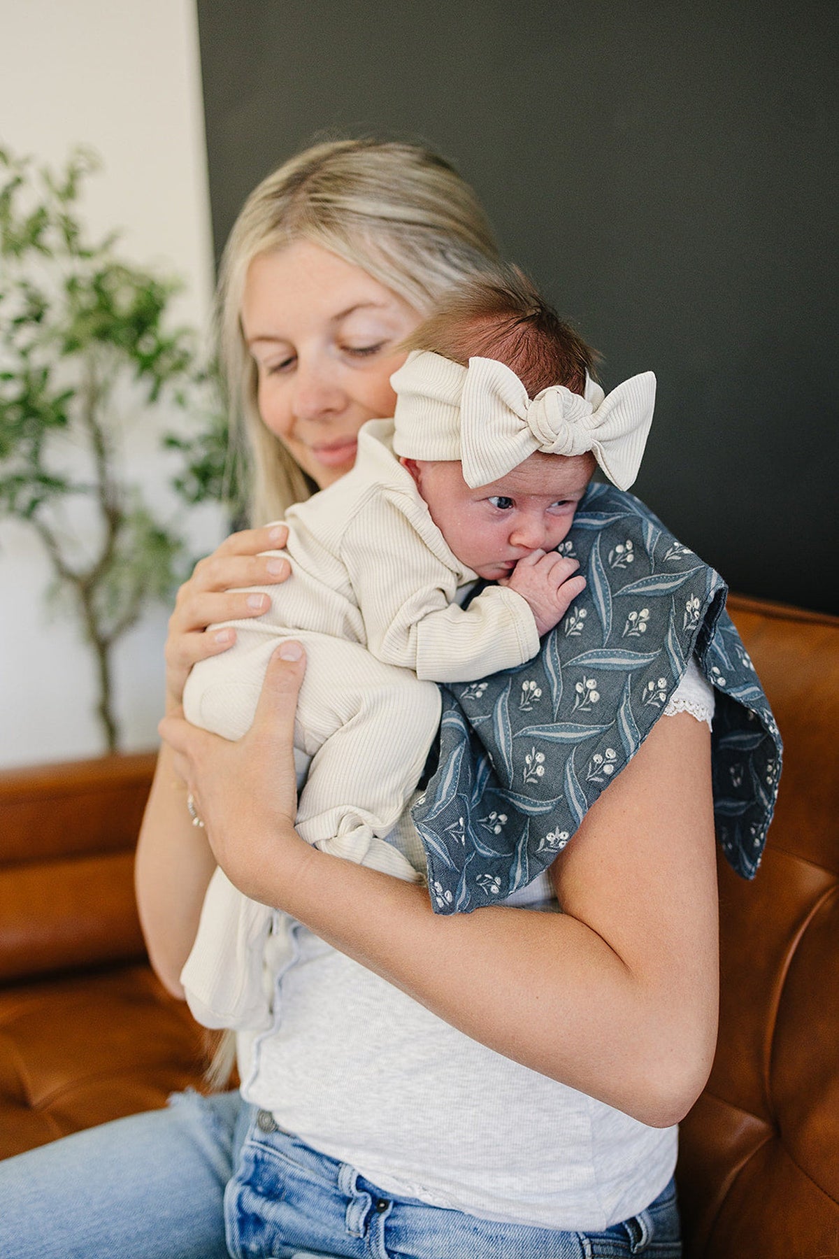 A woman holds a baby wearing a cream outfit and headband, resting on a navy floral muslin burp cloth.