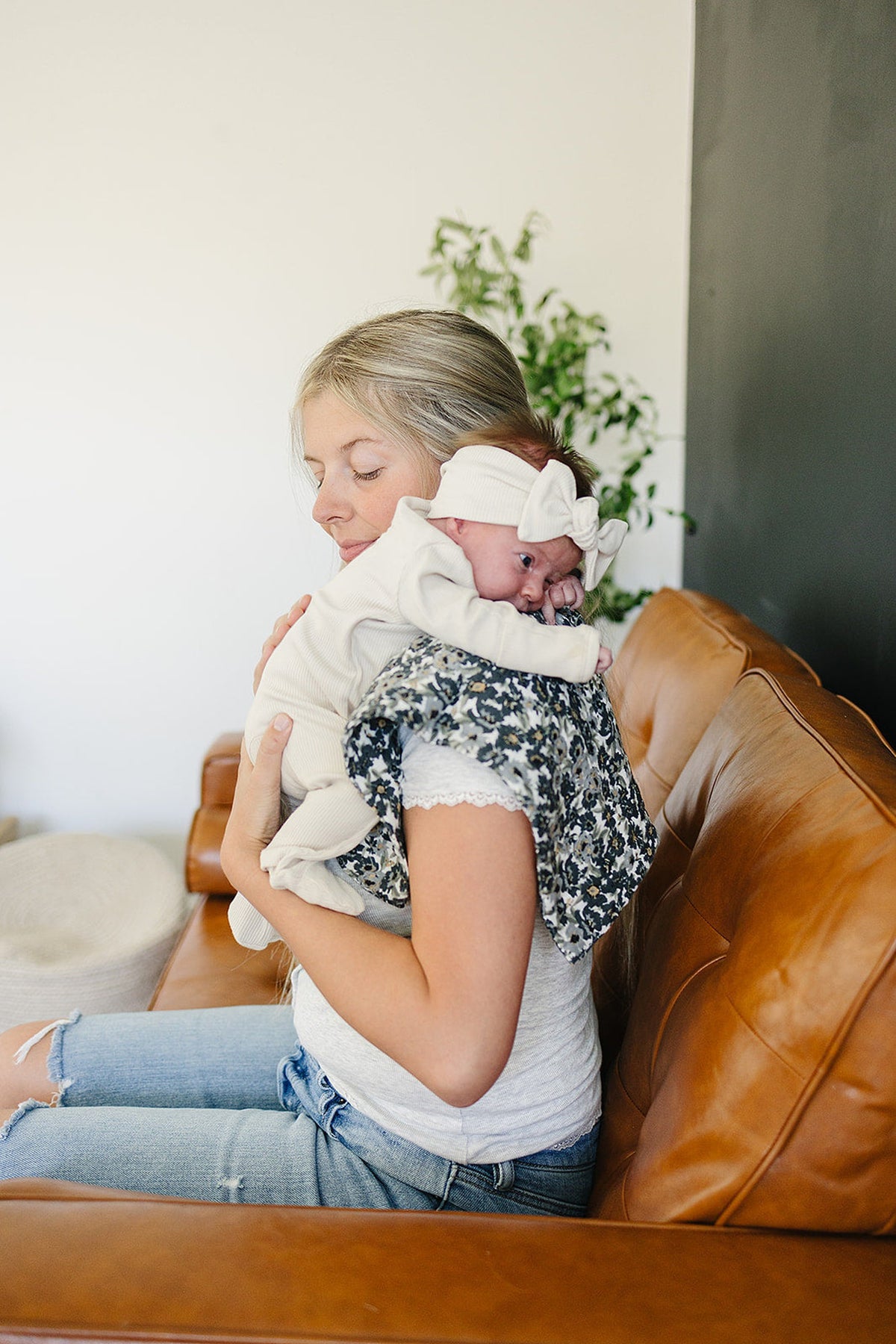 A woman holds a baby on her shoulder with a floral muslin burp cloth, sitting on a leather couch.