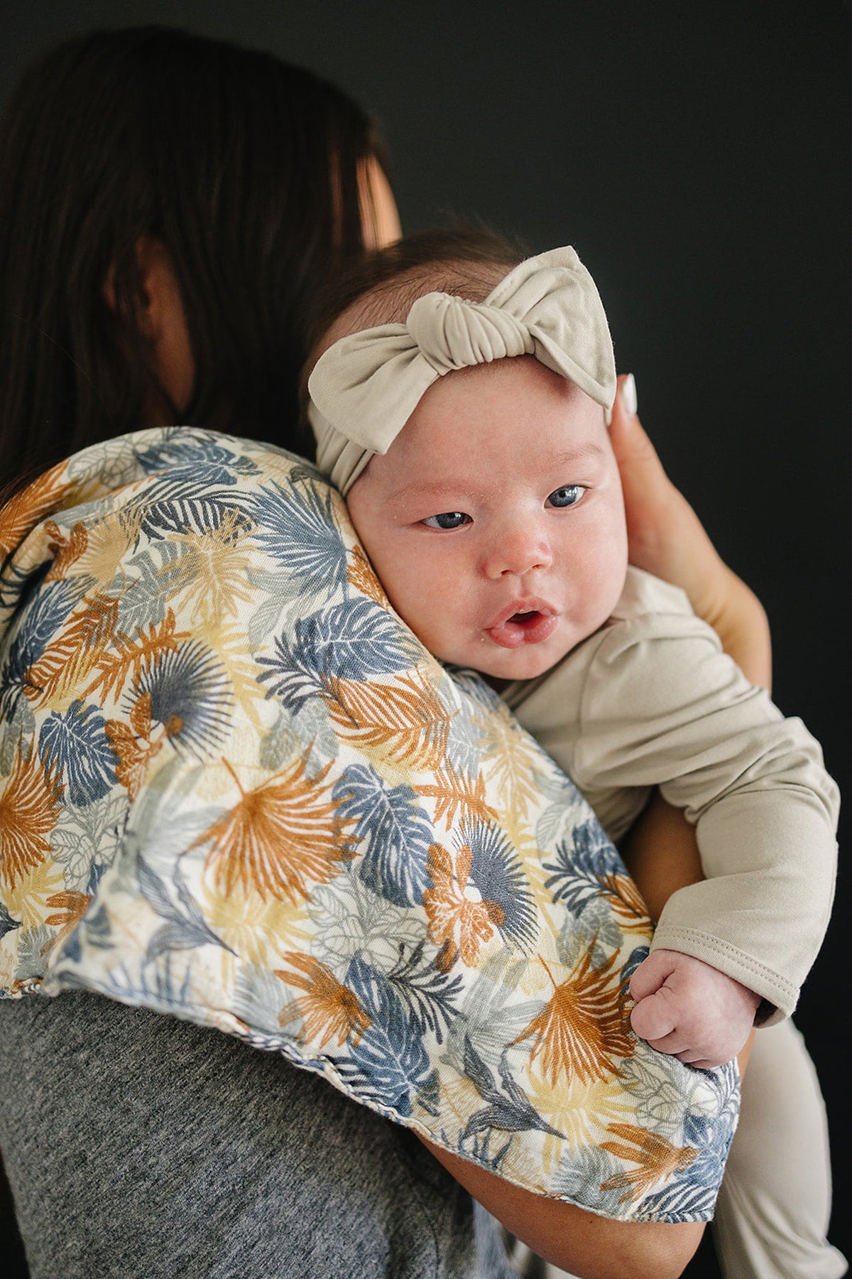A baby wearing a beige headband is held over a shoulder with a colorful palm-patterned muslin burp cloth.
