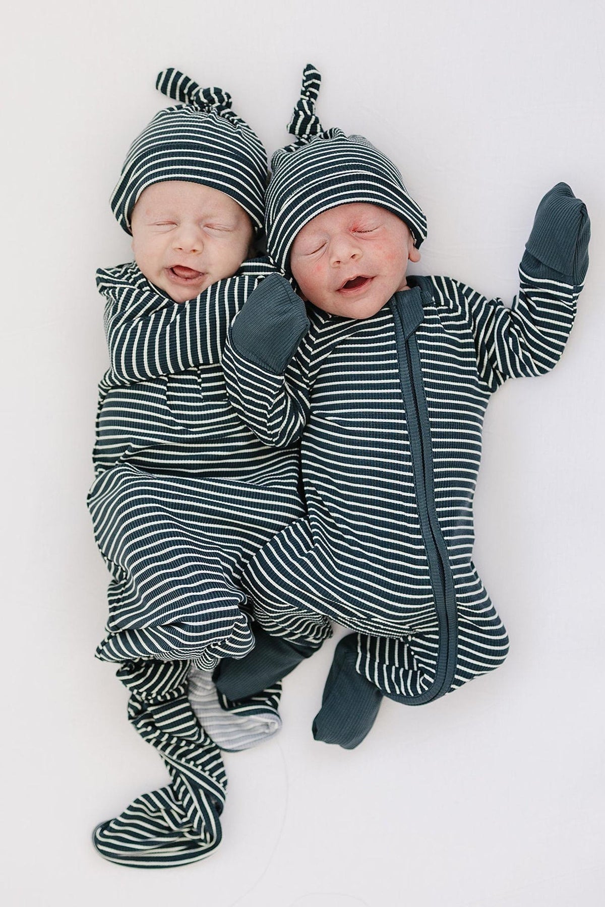 Two newborns in navy striped ribbed outfits and matching knot hats, lying on a soft white surface.
