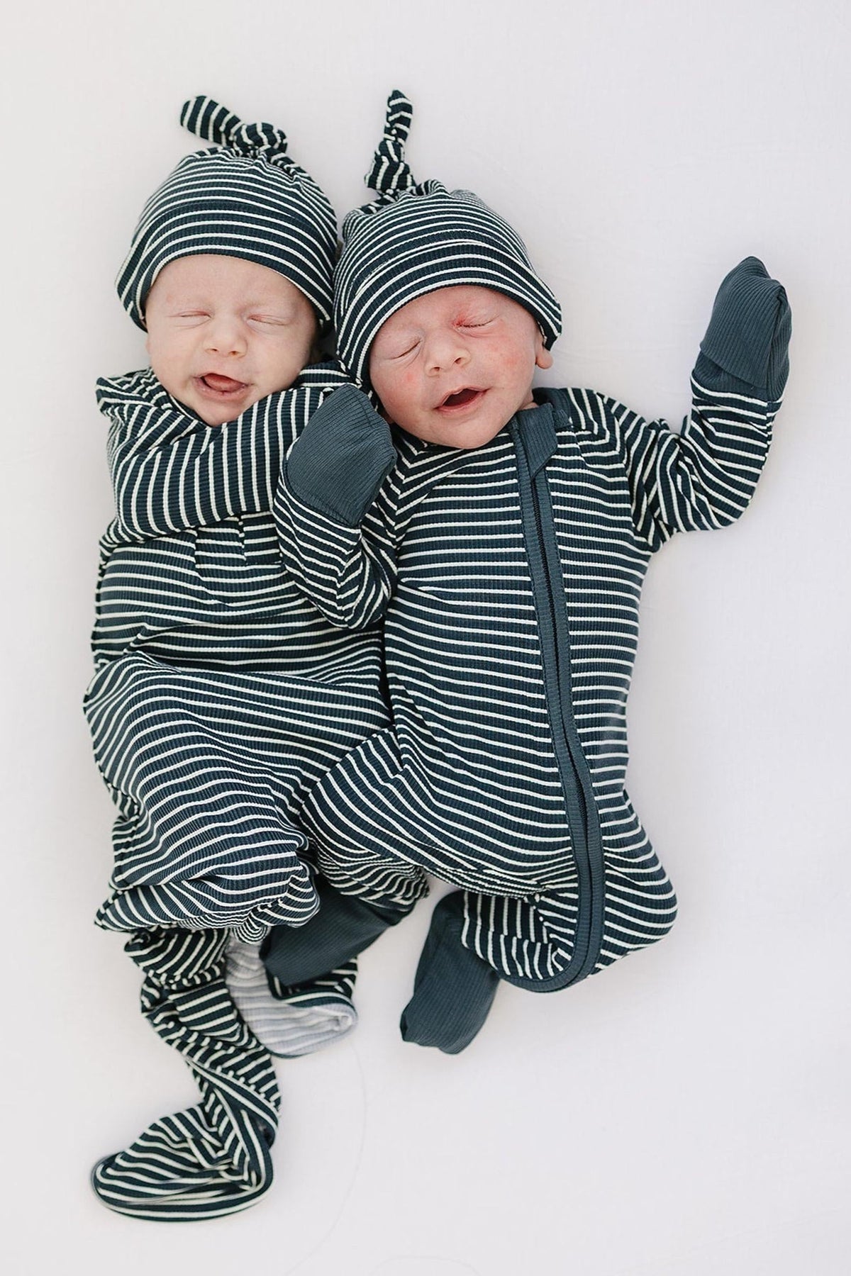 Two babies wearing navy and white striped pajamas with hats, lying on a white surface, smiling gently.