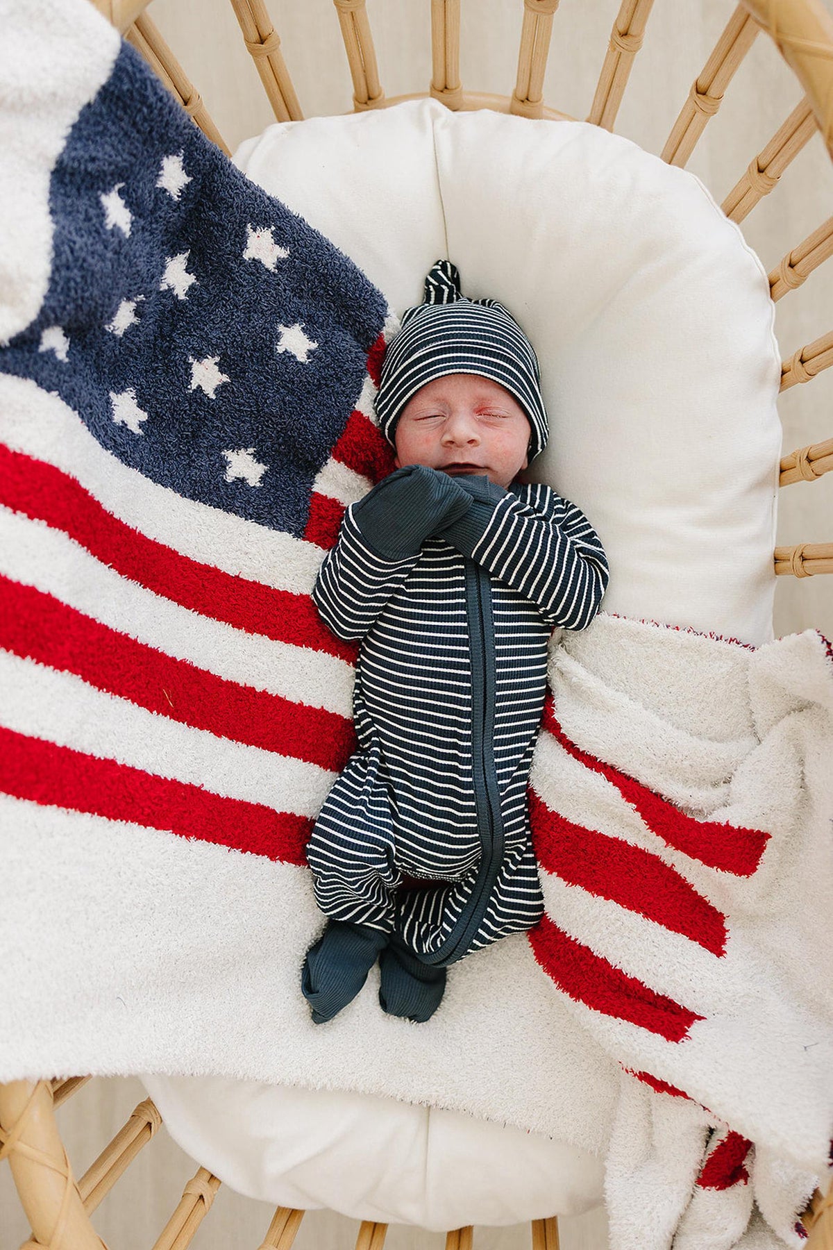 A baby in navy striped pajamas and hat sleeps on a cushion, wrapped in an American flag blanket.