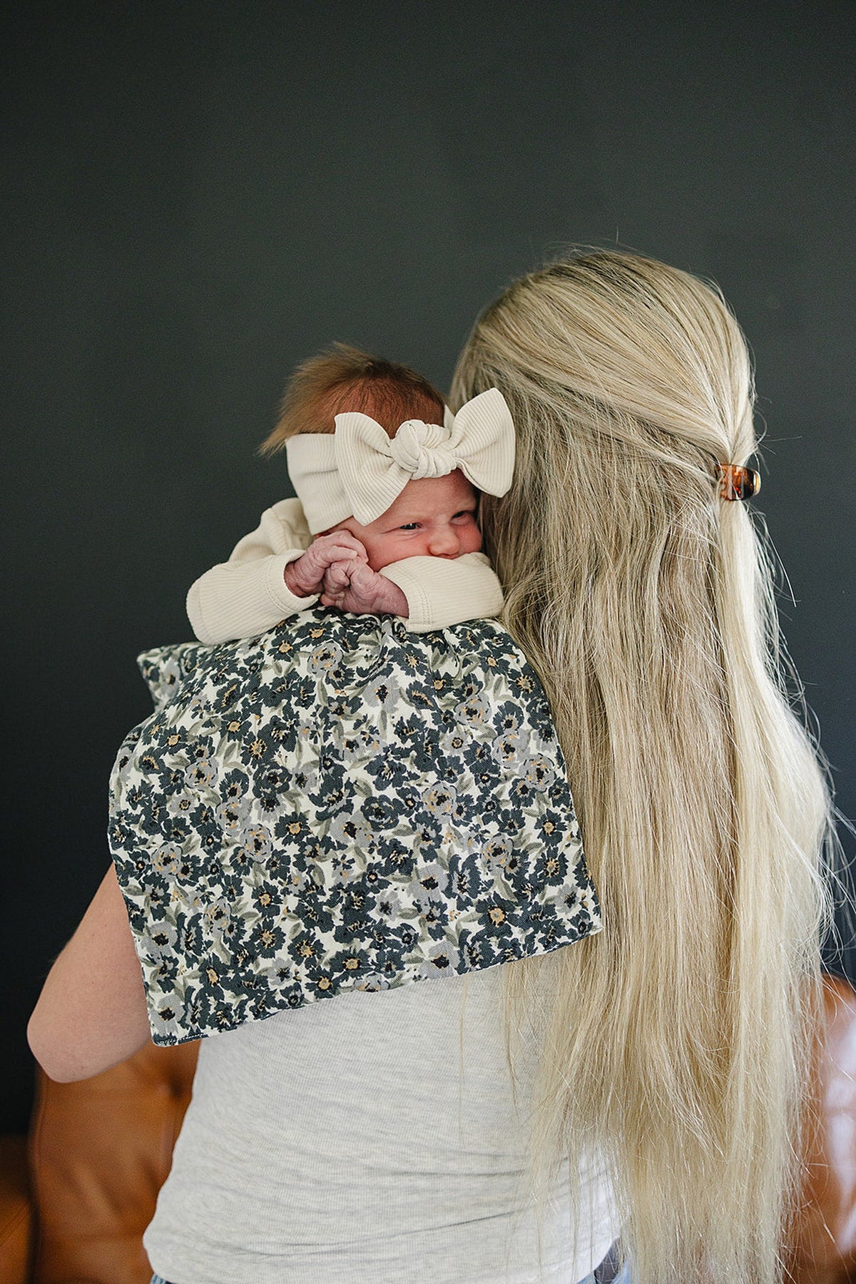 A baby with a cream bow headband rests on a floral muslin burp cloth draped over a womans shoulder.