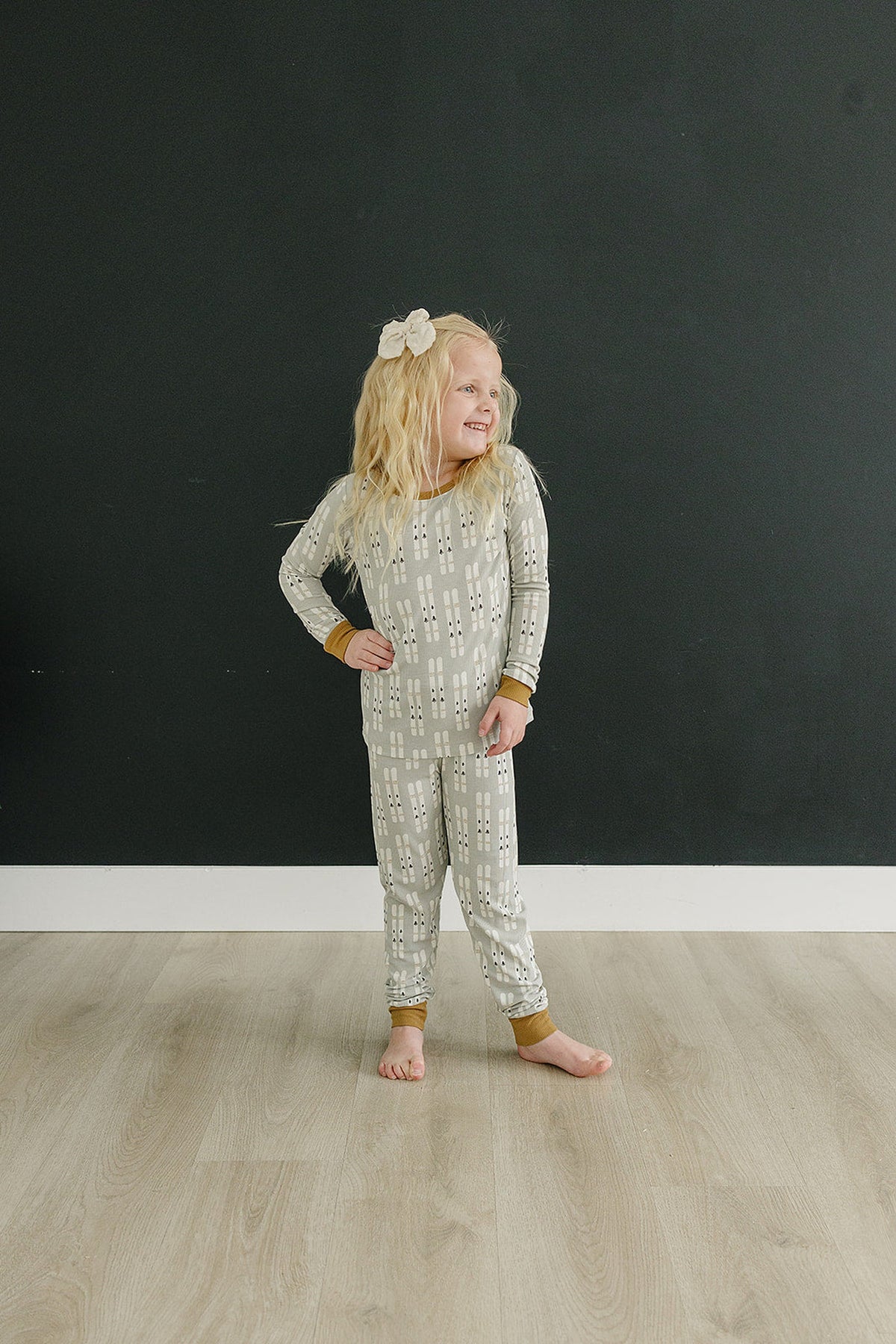 A child wearing a gray, ski-patterned pajama set with brown cuffs, standing on a light wooden floor.
