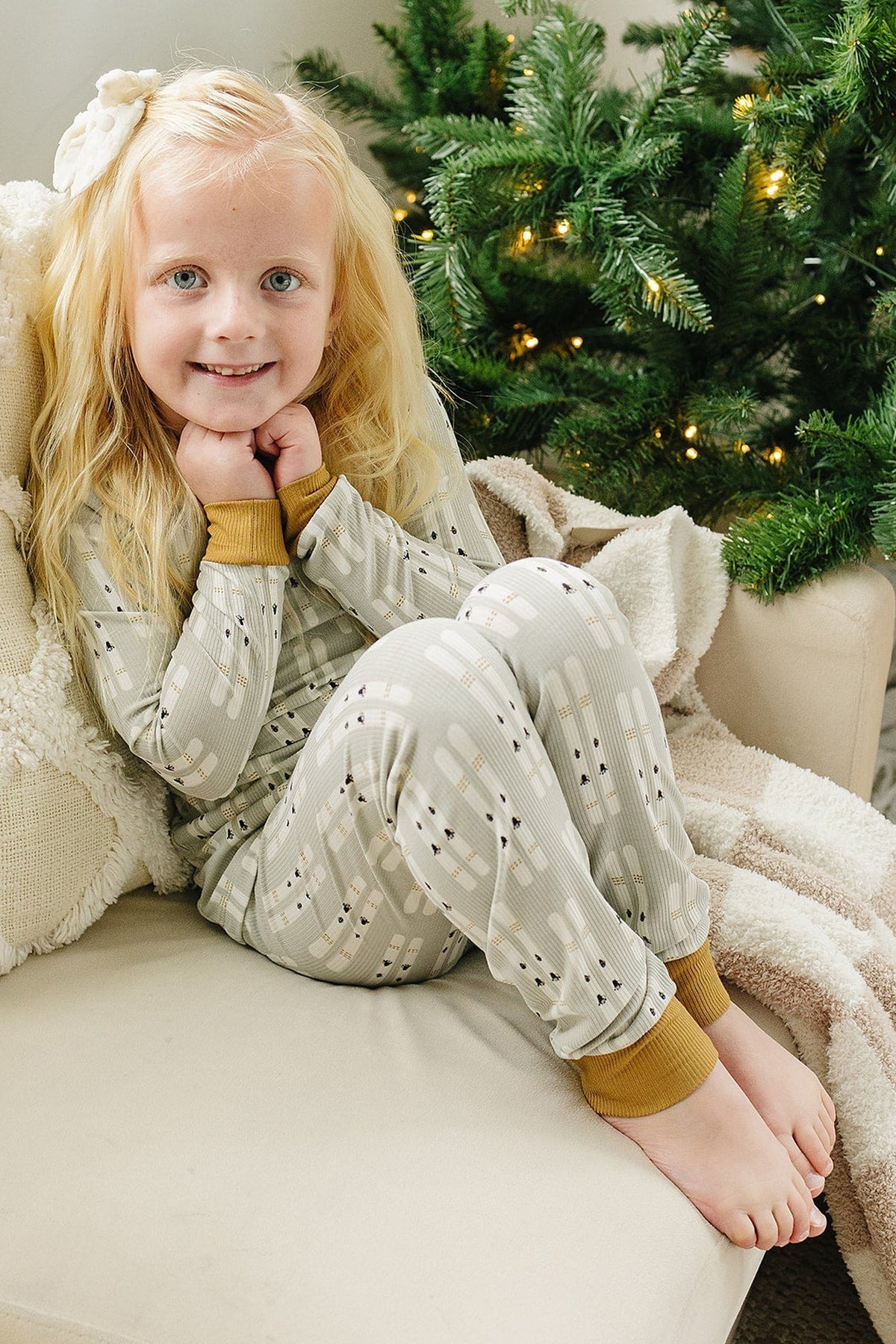 A child in ski-themed pajamas sits on a couch beside a Christmas tree, smiling warmly.