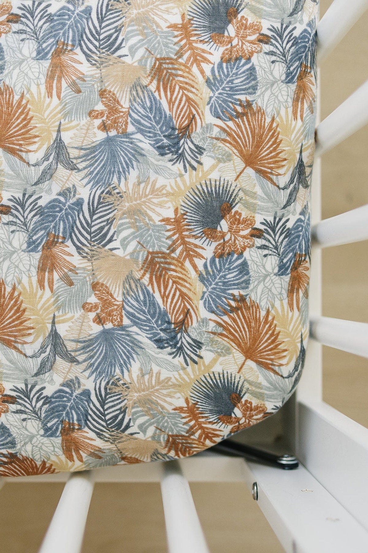 Tropical leaf-patterned muslin crib sheet in blue, brown, and beige hues on a white crib.