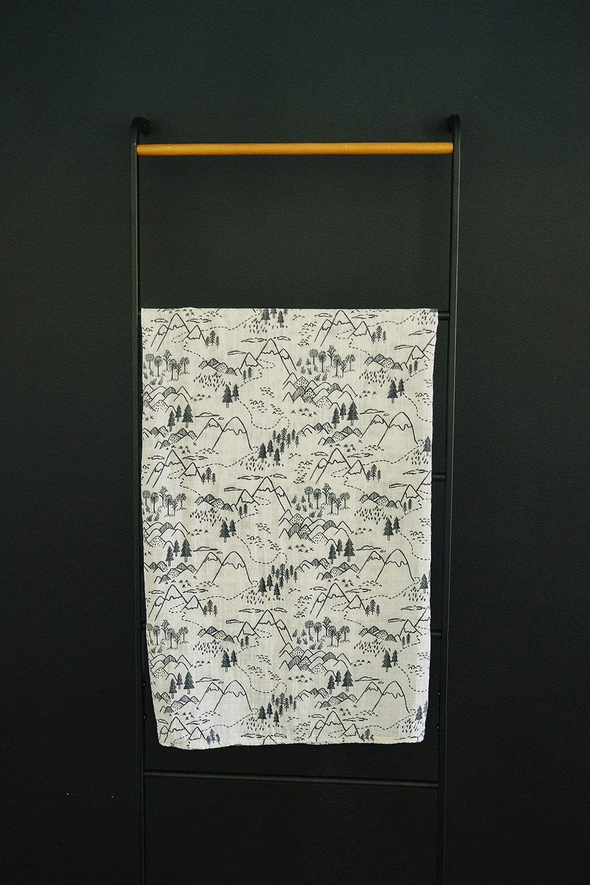 White muslin swaddle blanket with black mountain and tree pattern on a black metal rack against a dark wall.