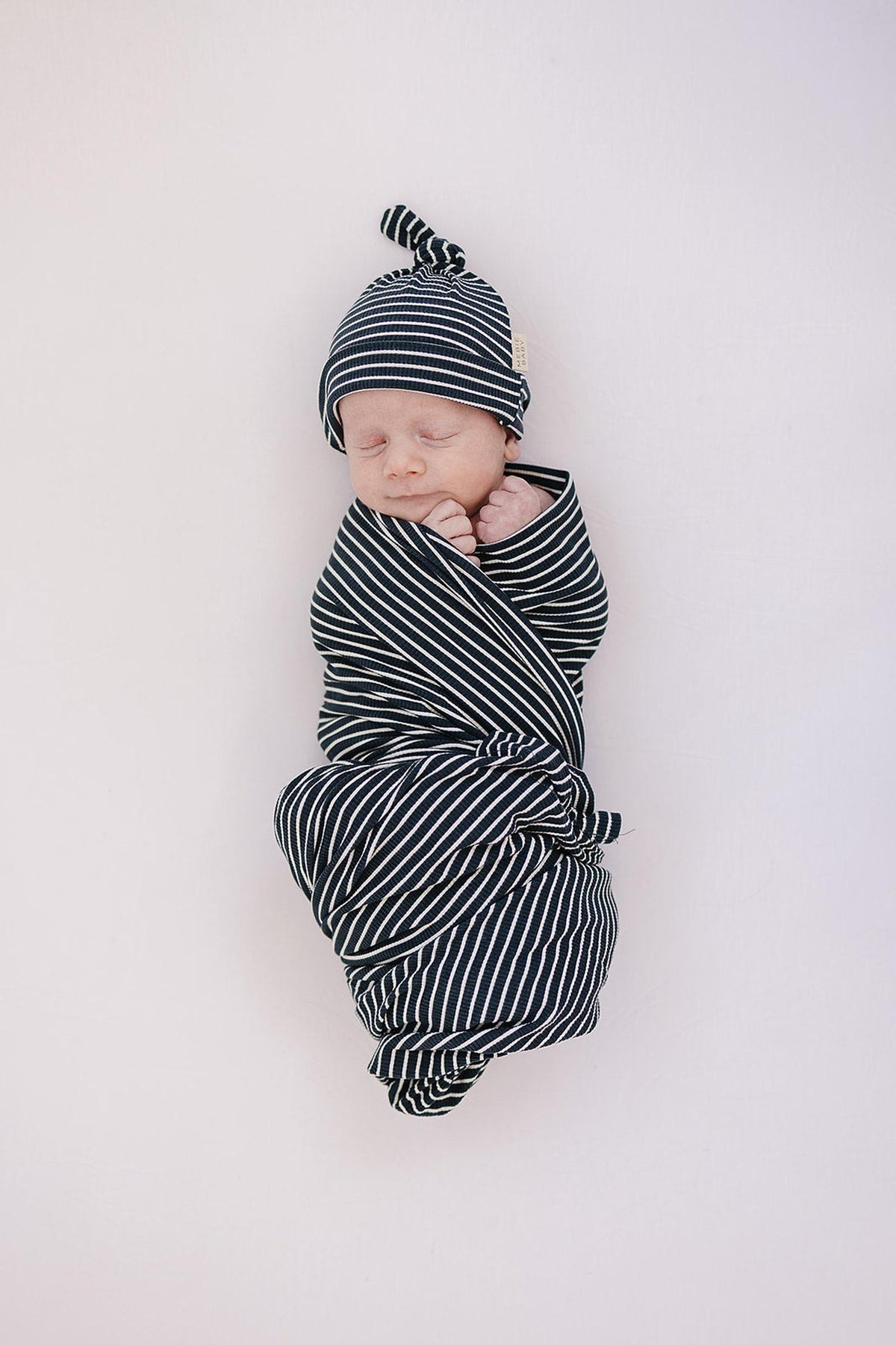 A newborn baby is wrapped in a navy and white striped swaddle with a matching knot hat, sleeping peacefully.