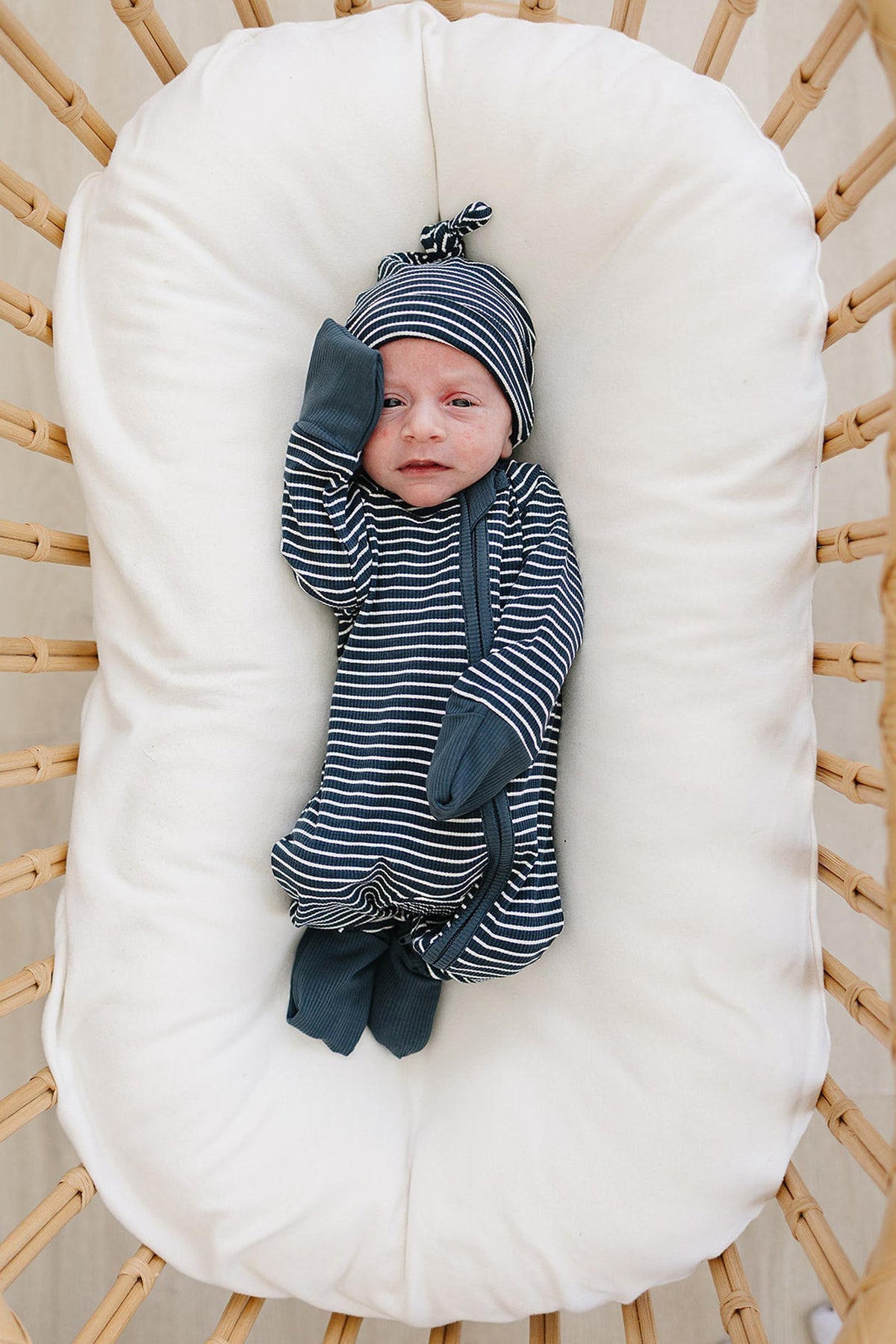 Newborn in navy striped bamboo pajamas and hat on a white cushion in a wicker bassinet.