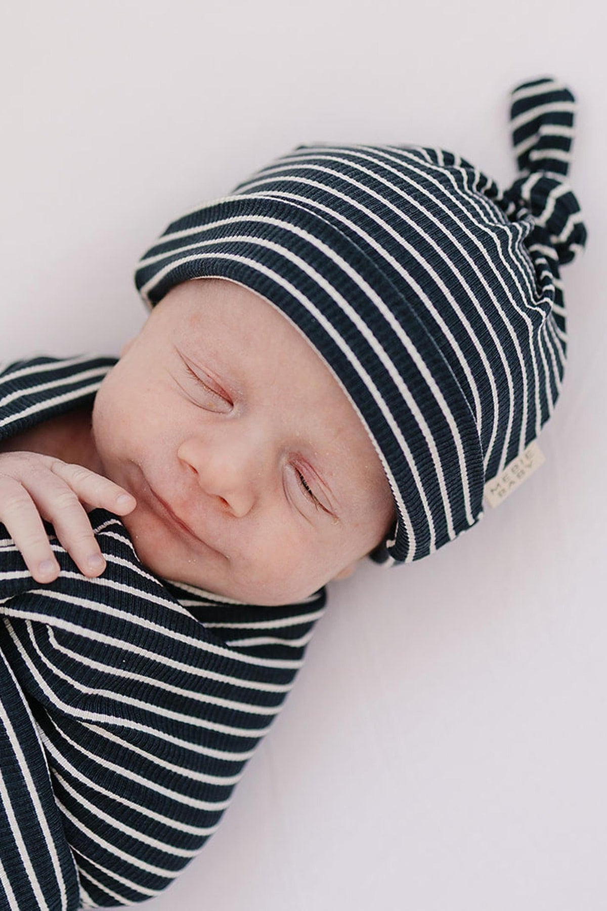 Newborn wearing a navy and white striped knot hat and matching swaddle, peacefully sleeping on a white background.