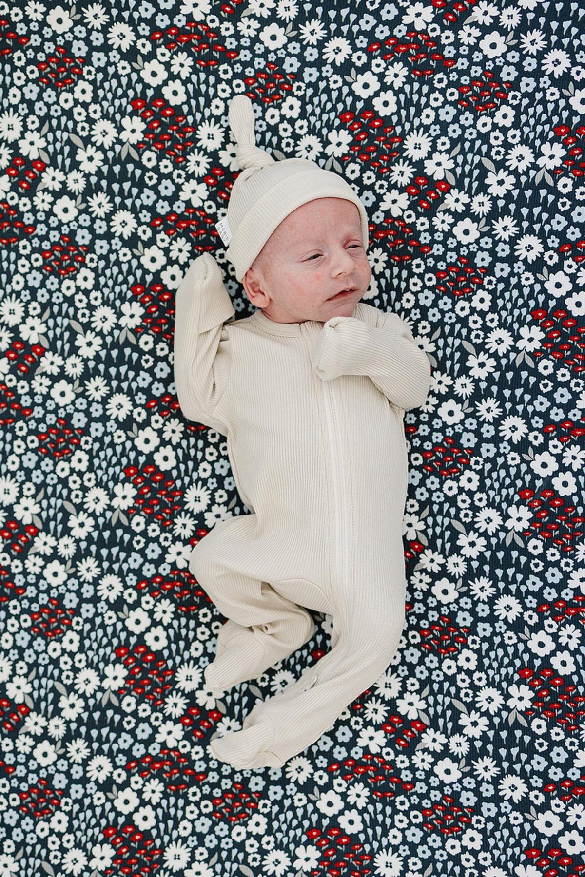 A baby in a cream outfit and hat lies on a floral-patterned crib sheet with white and red flowers.