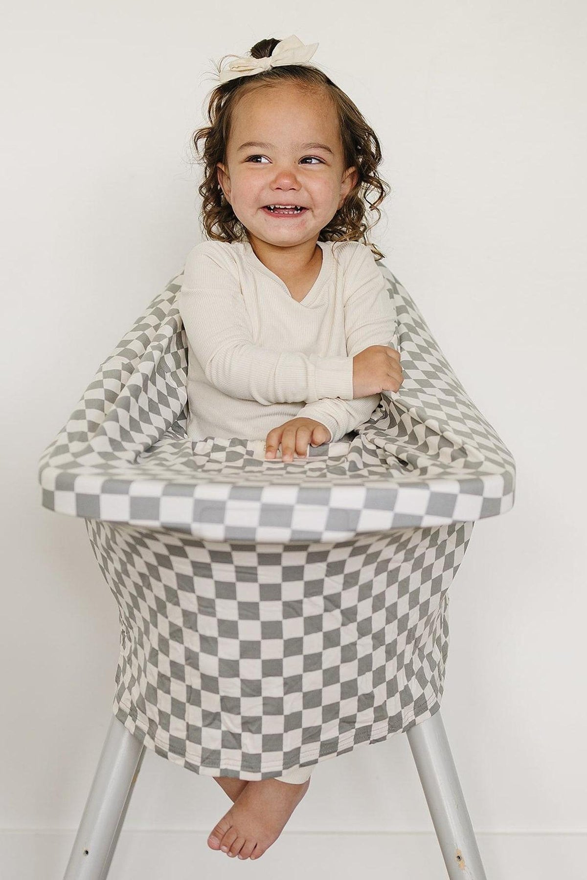 A smiling child sits in a high chair covered with a light green checkered fabric, wearing a cream-colored outfit.