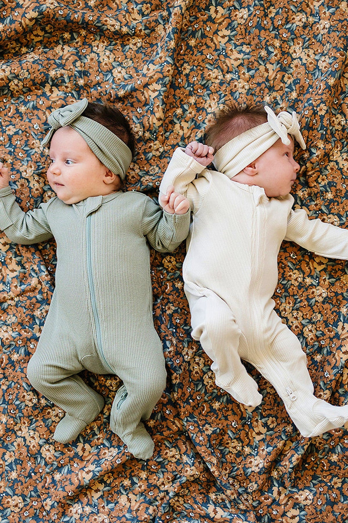 Two babies wearing ribbed onesies and headbands lie on a floral-patterned quilt, one in green, one in cream.