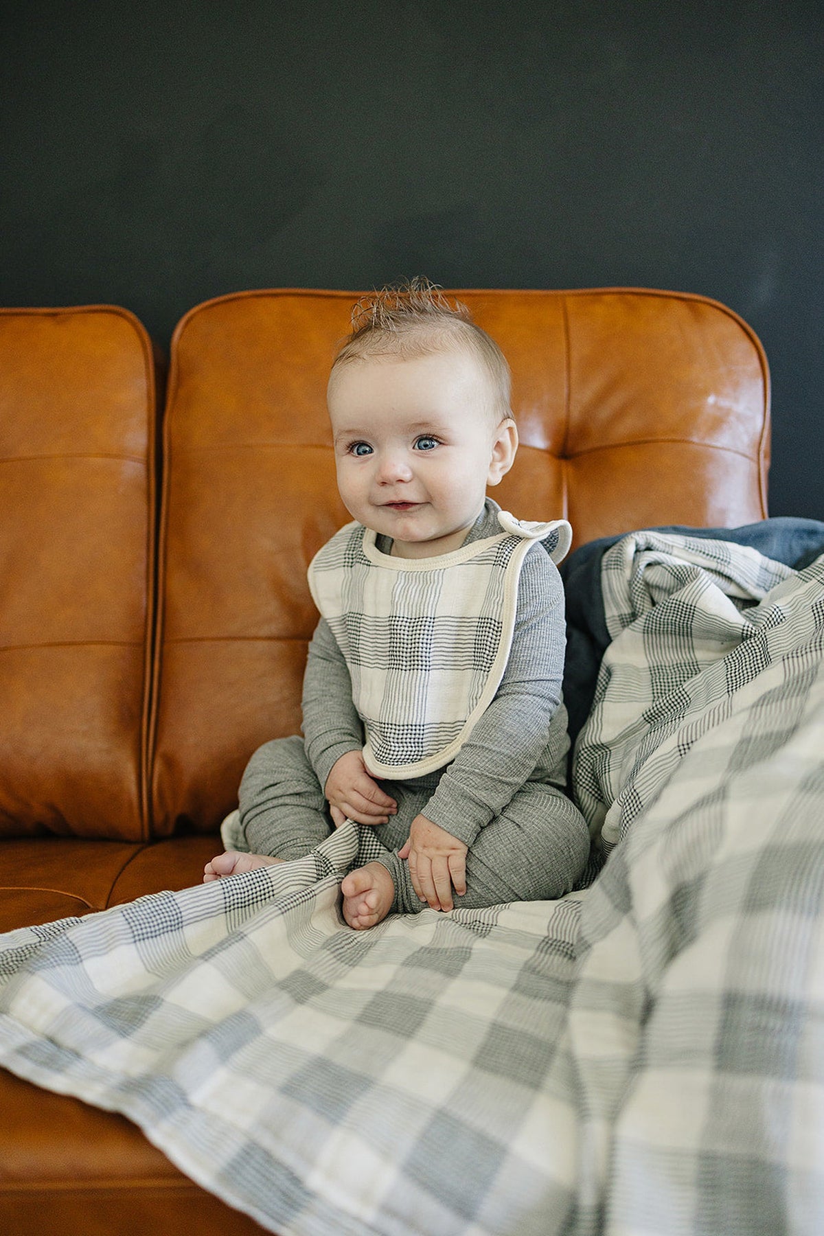 A baby wearing a plaid bib sits on a brown leather couch with a matching plaid quilt.