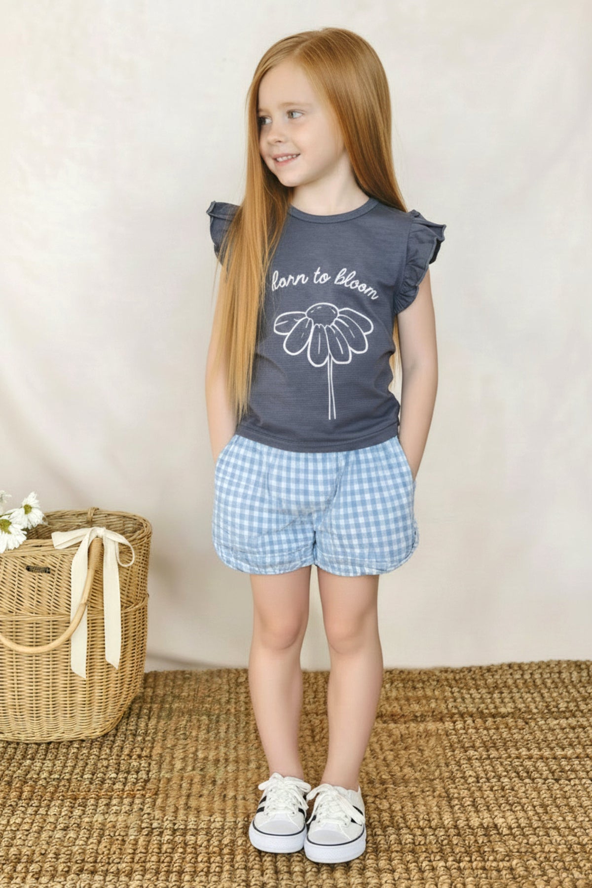 A young girl with long red hair wears a navy blue shirt with a floral design and blue gingham shorts.