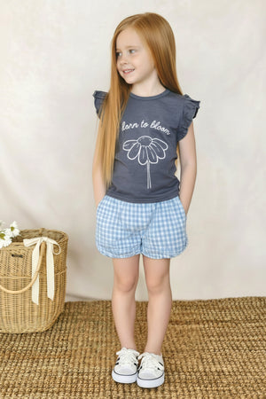 A young girl with long red hair wears a navy blue shirt with a floral design and blue gingham shorts.