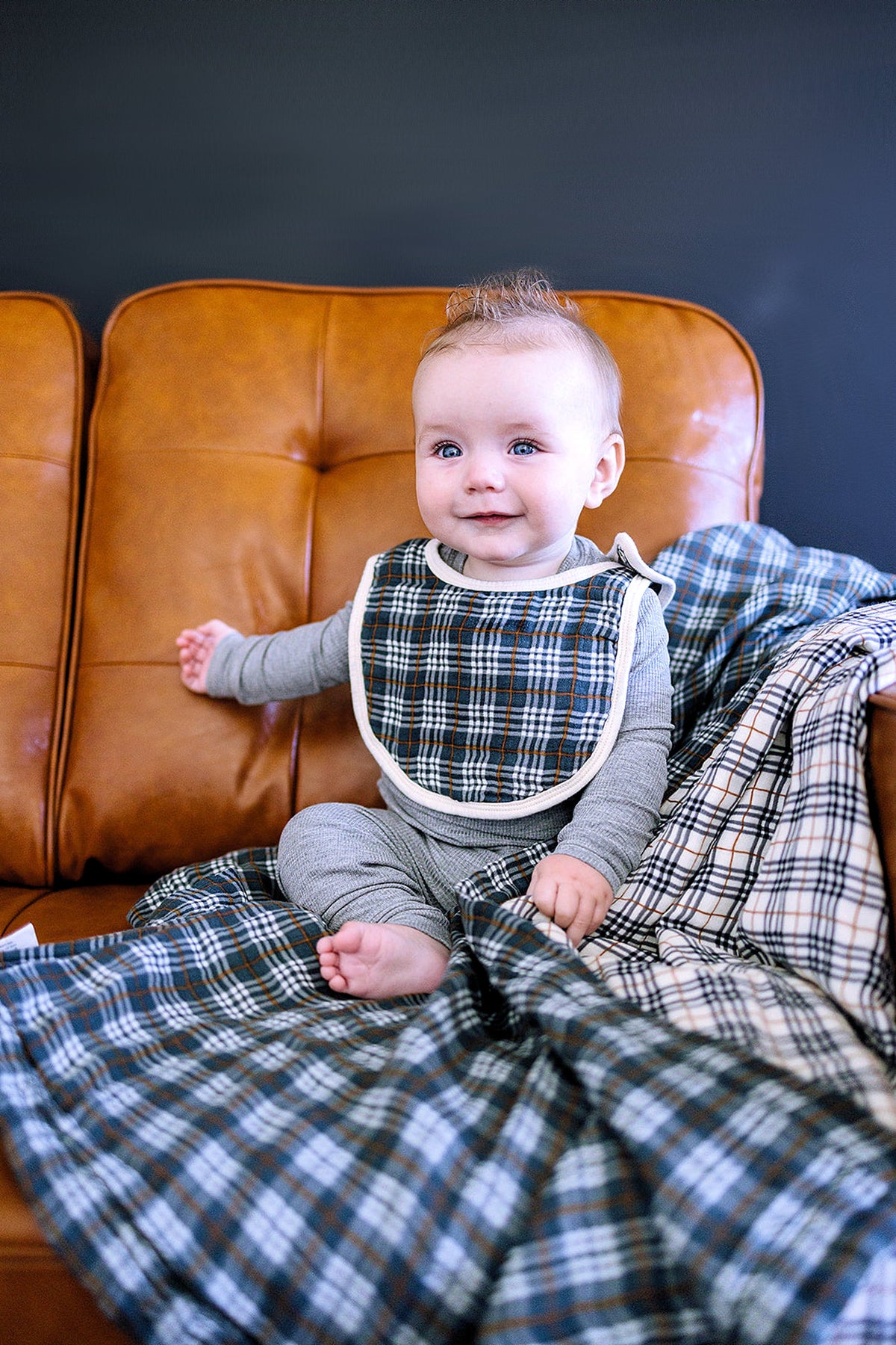 A baby wearing a navy plaid muslin bib sits on a brown leather couch with matching plaid blanket.
