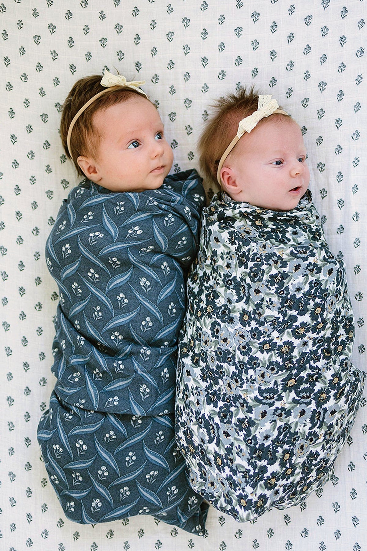 Two babies swaddled in patterned muslin blankets, one navy with floral designs and the other white with floral prints.