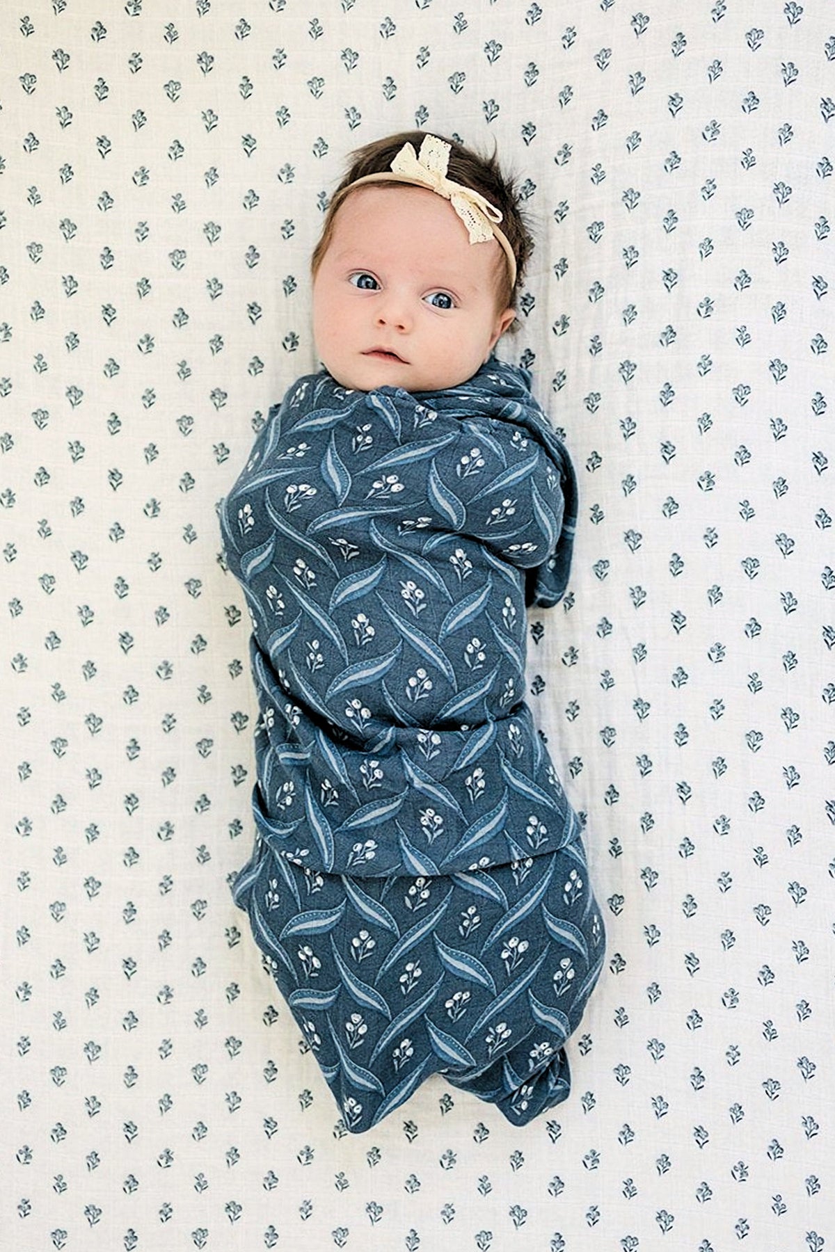 A baby is wrapped in a navy patterned muslin swaddle, lying on a matching floral blanket, wearing a headband.