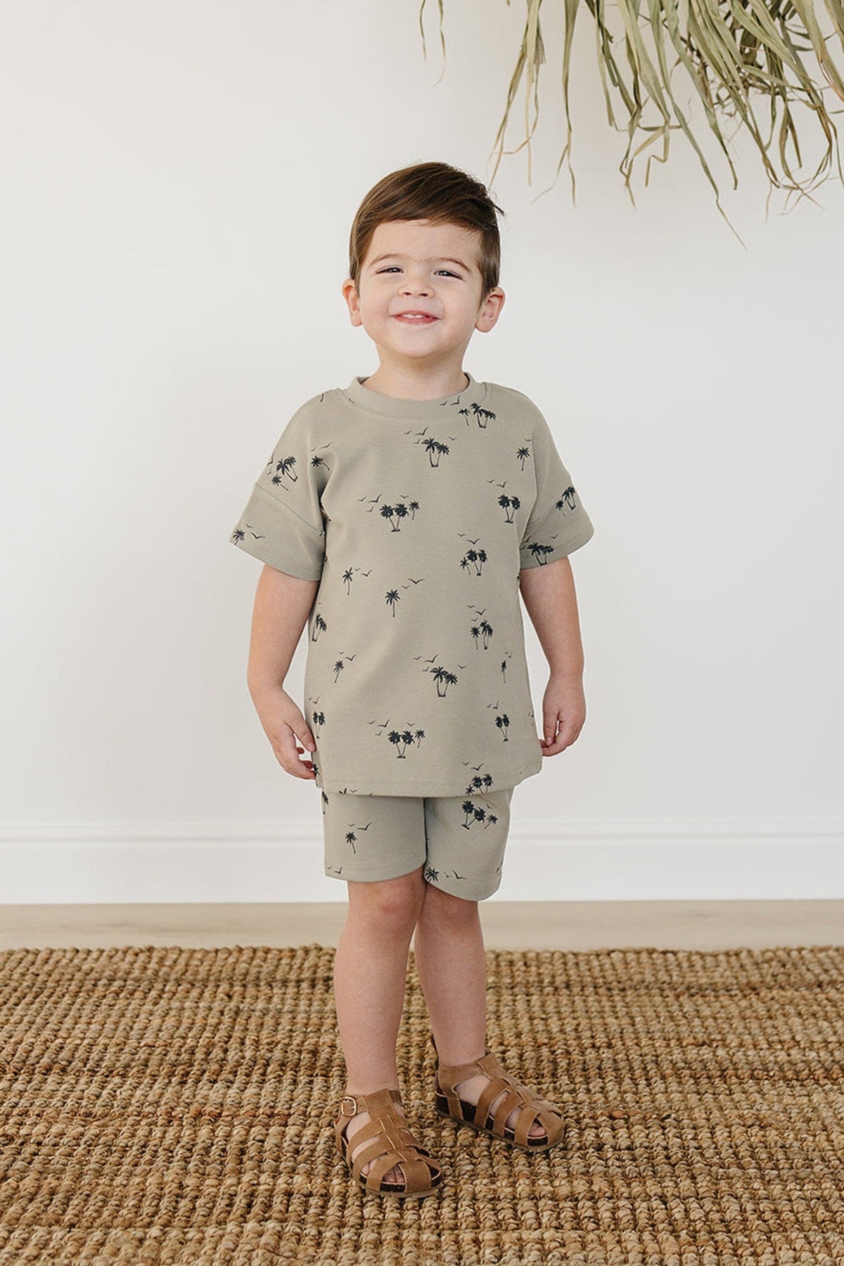 A young child smiles while wearing a light green palm tree print short sleeve shirt and matching shorts. The outfit is made of soft cotton, perfect for toddlers.