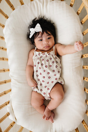 A baby girl rests on a soft, light-colored cushion, wearing a floral-patterned romper and a white bow in her hair.