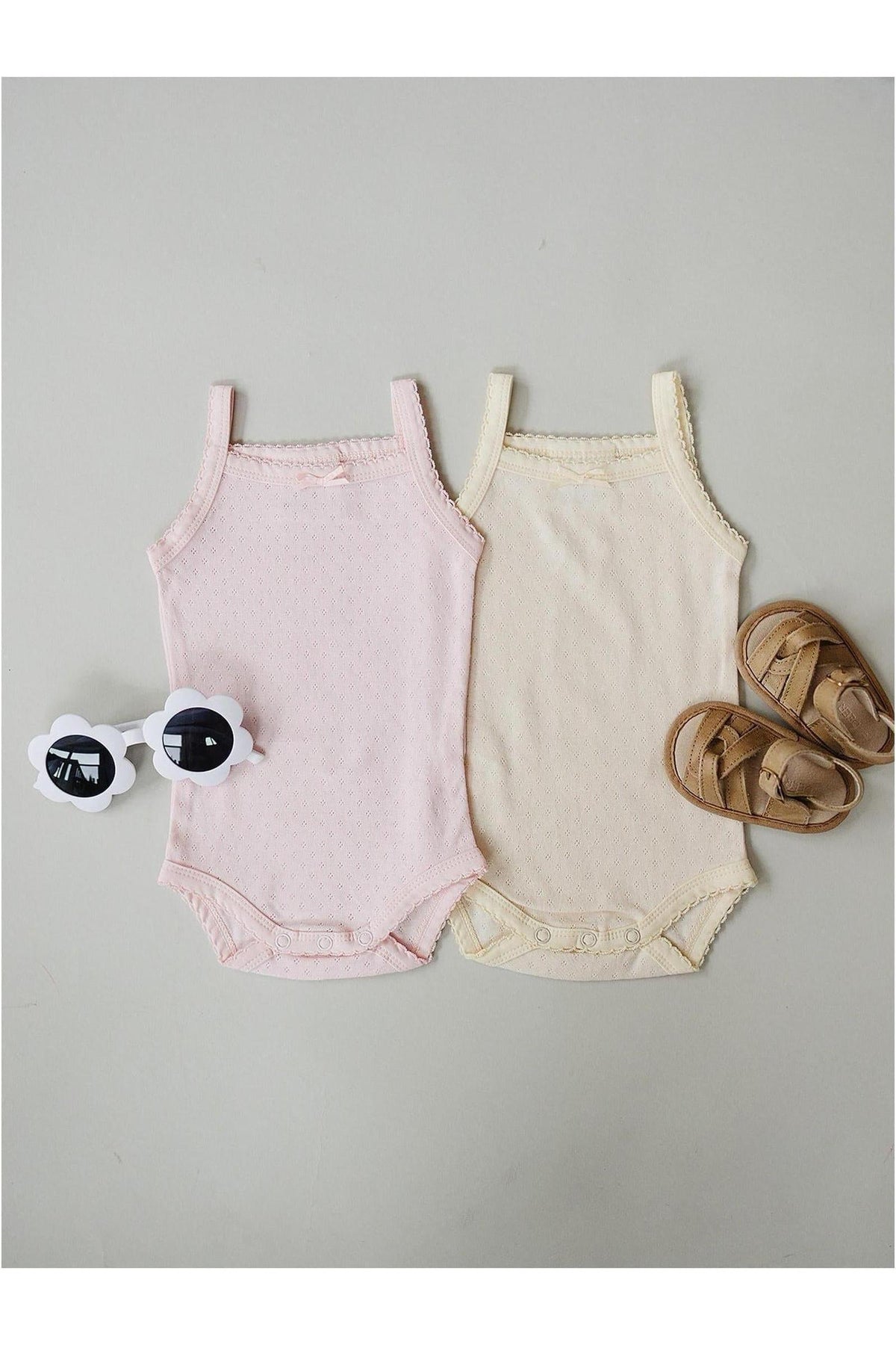 Two baby tank bodysuits in pink and yellow cotton are displayed alongside a pair of brown sandals and white sunglasses.