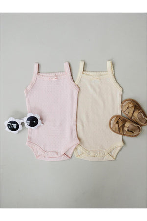Two baby tank bodysuits in pink and yellow cotton are displayed alongside a pair of brown sandals and white sunglasses.
