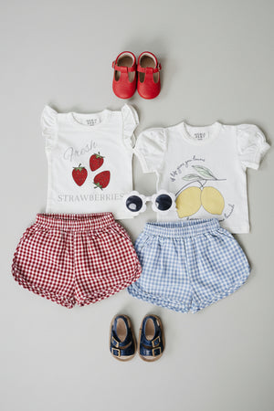 Flat lay of two pairs of shorts, one red gingham and one blue gingham, paired with matching graphic t-shirts and accessories, showcasing a playful summer outfit for kids.