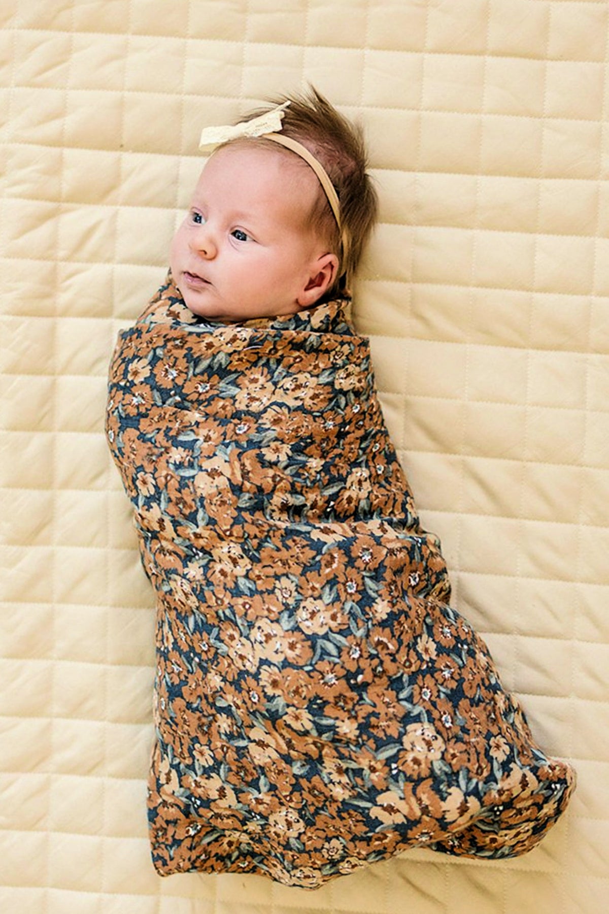 Baby wrapped in a floral-patterned swaddle blanket lies on a quilted cream surface, wearing a headband.