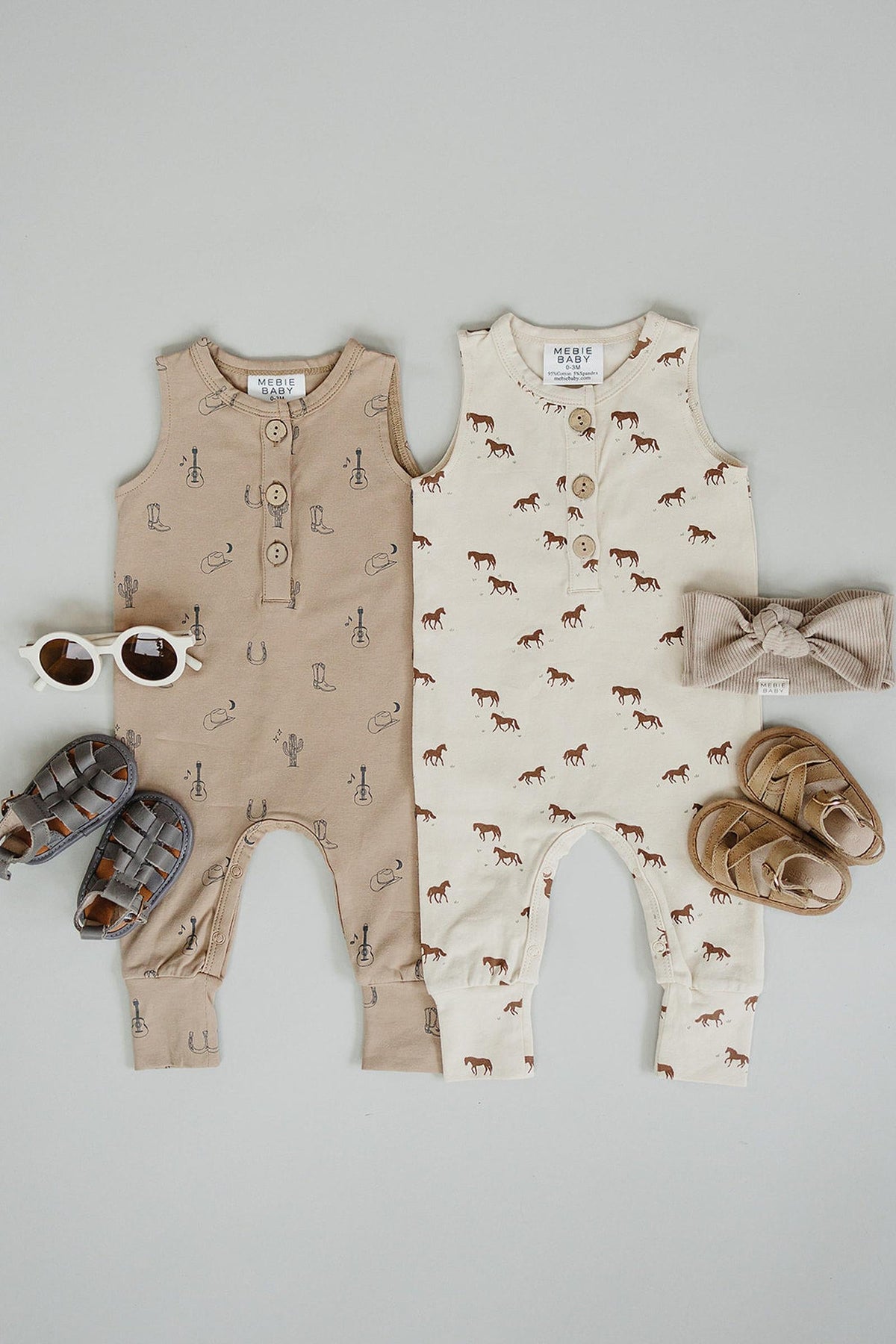 Two baby tank rompers in beige and cream, featuring playful horse graphics, paired with sandals and sunglasses on a light.