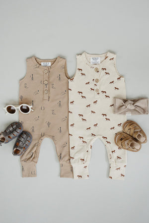 Two baby tank rompers in beige and cream, featuring playful horse graphics, paired with sandals and sunglasses on a light.